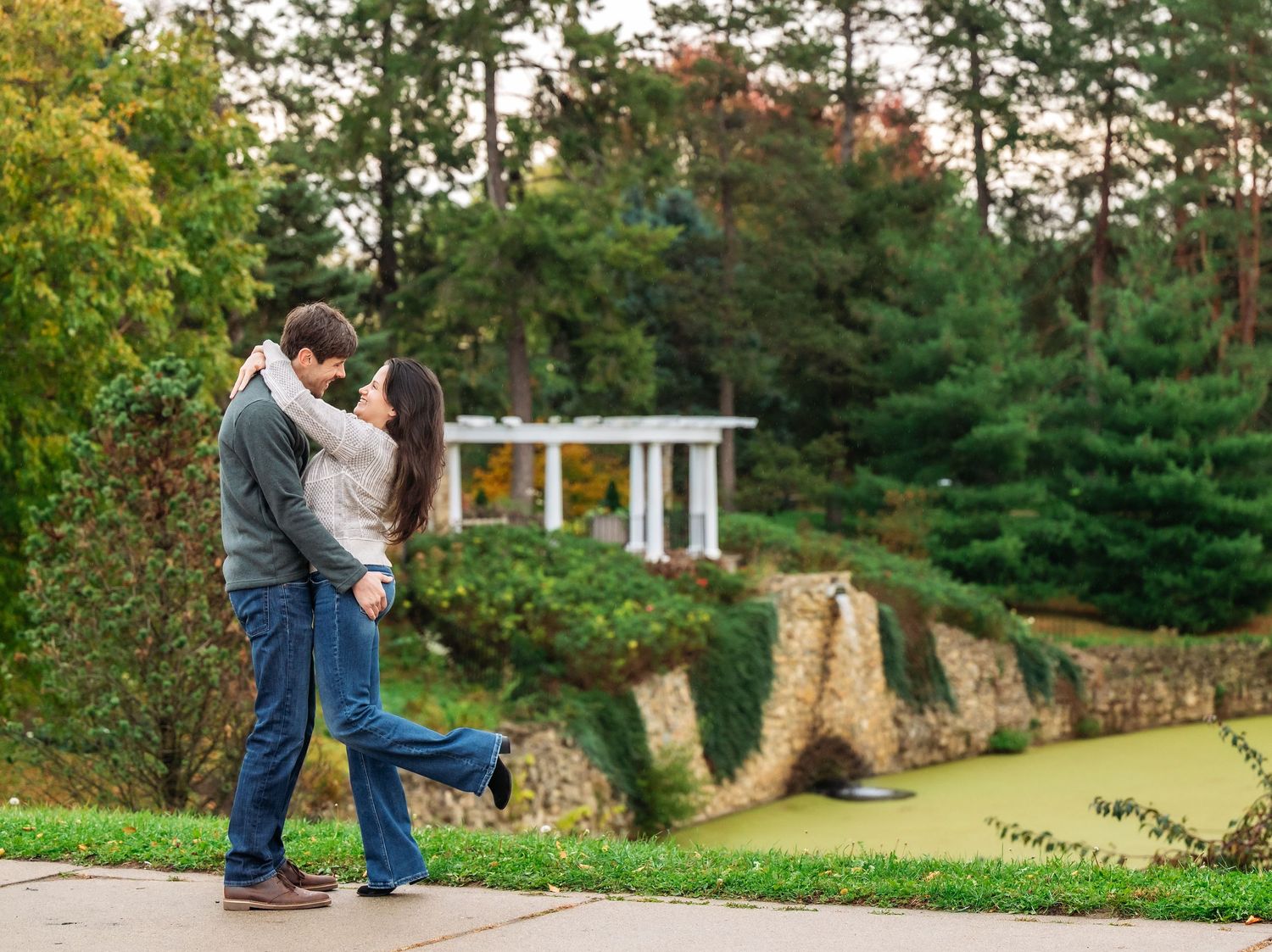 A romantic dance sequence unfolds in a park with stone walls and evergreen trees in the background.