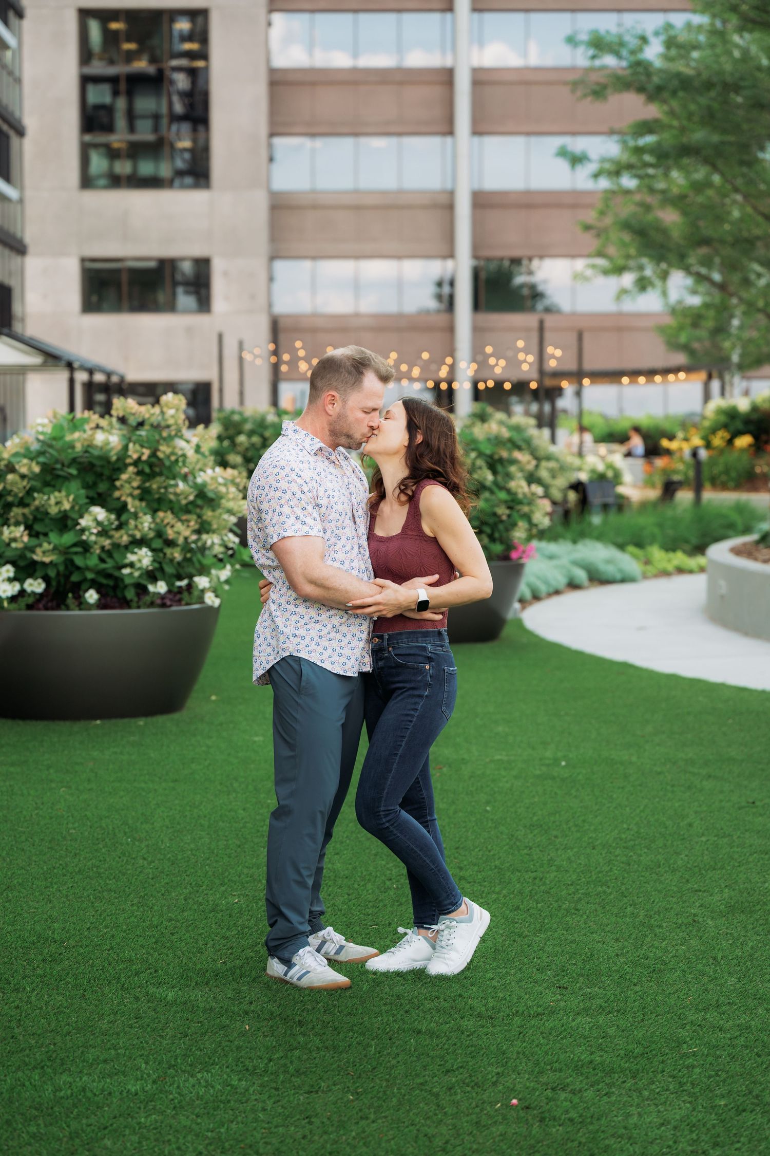 A couple shares an intimate dance moment on a green lawn with flowering bushes and a building in the background.