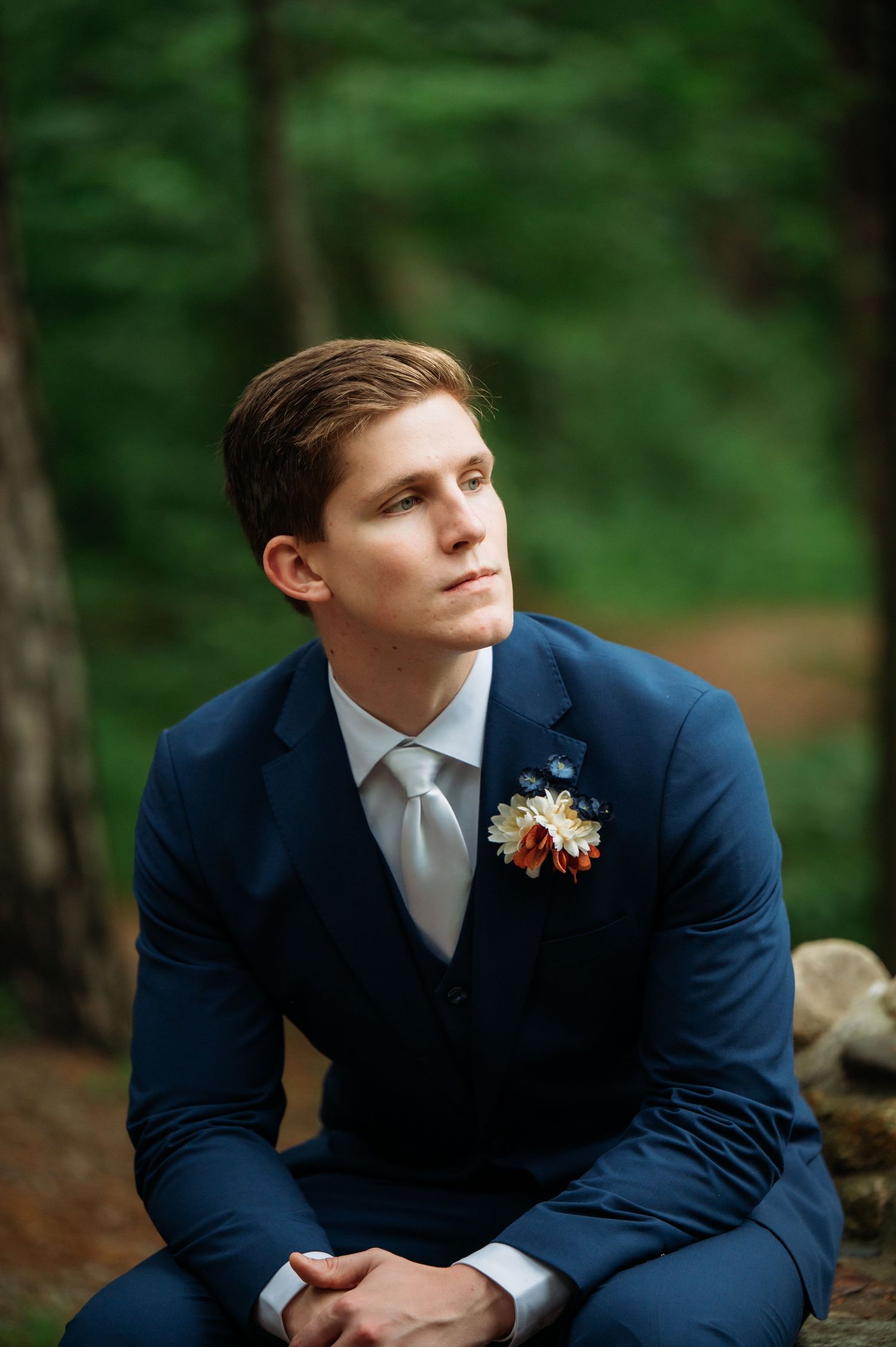 A groom in a navy blue suit with boutonniere sits thoughtfully in a forest setting.