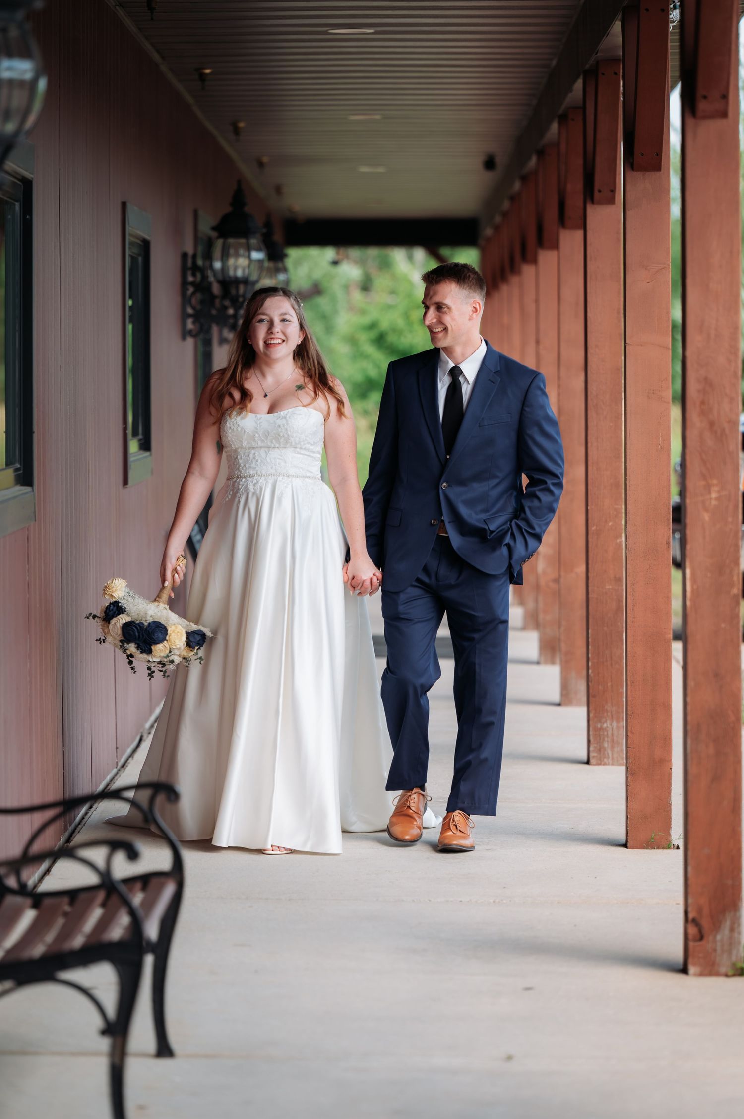 Couple in wedding attire walking and holding hands along a covered outdoor walkway with wooden posts at Northern Lights near Milaca MN..