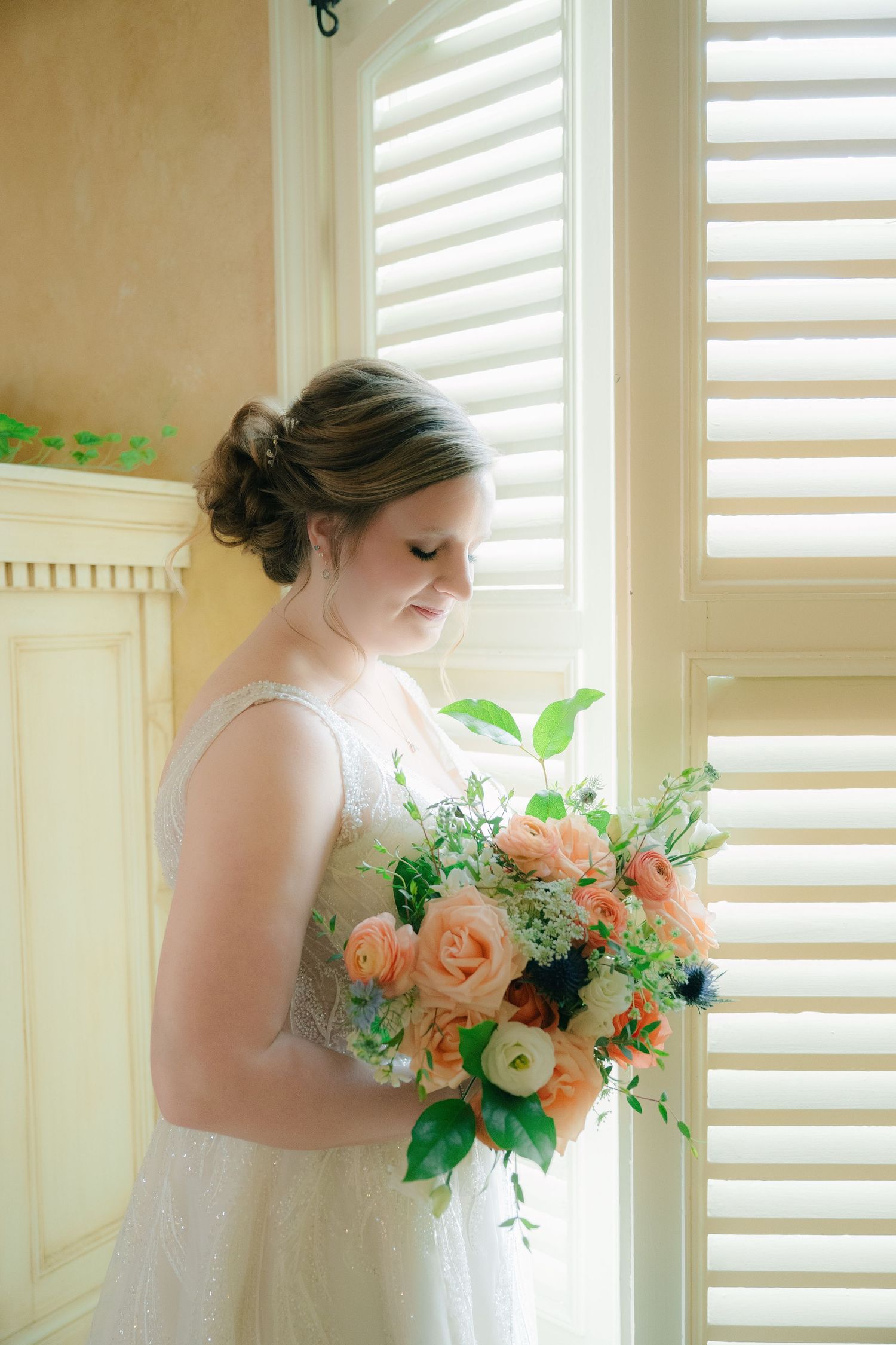 Bride in white lace gown holds peach and green bouquet by shuttered window.