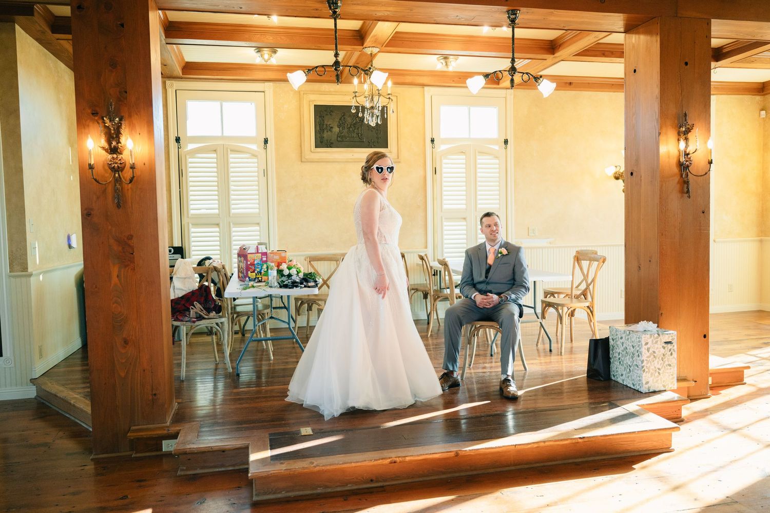 Bride in white gown with sunglasses stands on steps while groom sits in chair at rustic wedding venue.