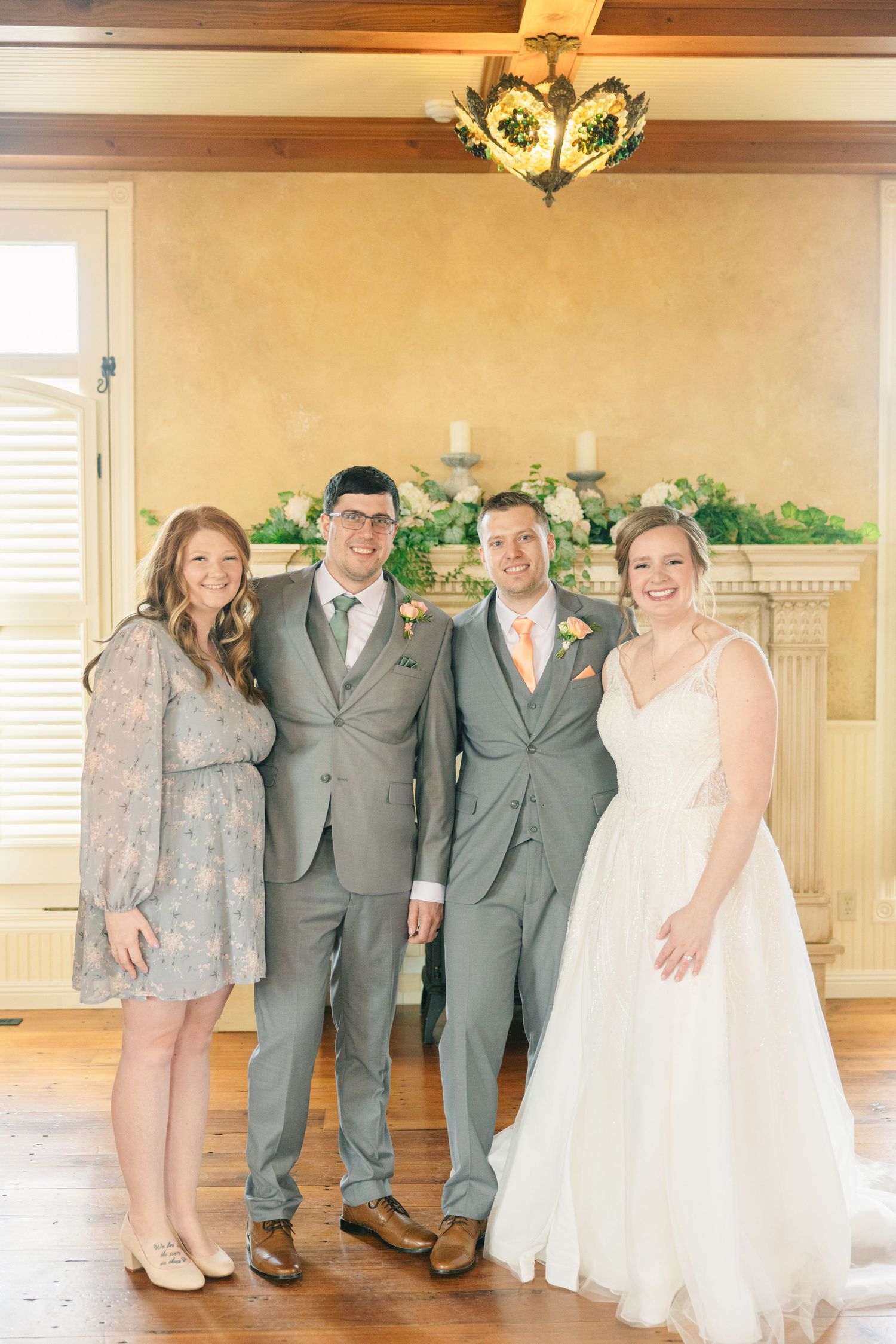 Wedding party group photo with bride in white gown, groom and groomsman in gray suits, beside a fireplace.