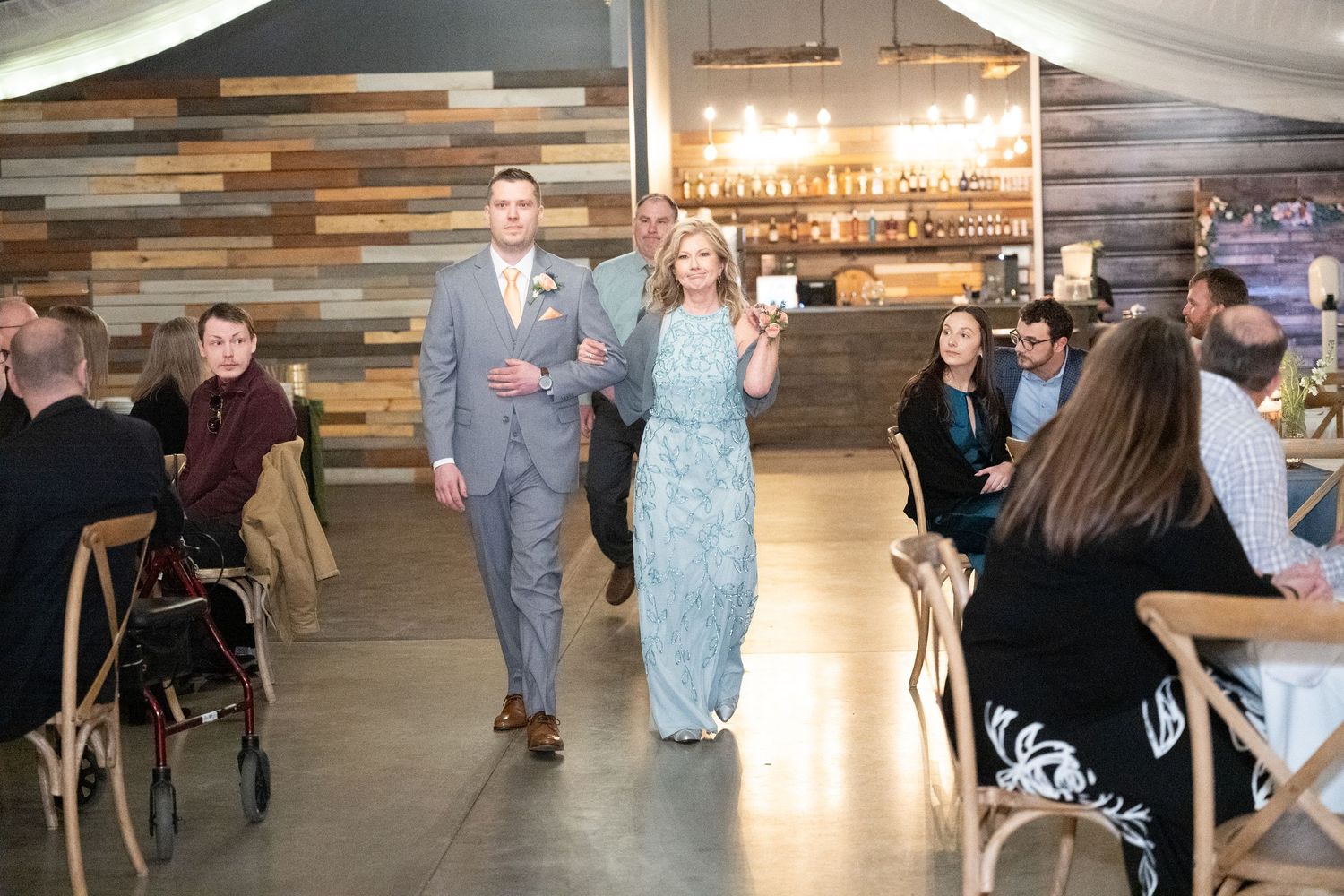 Groom escorts a woman in blue down the aisle at a rustic indoor wedding venue with wood accents.