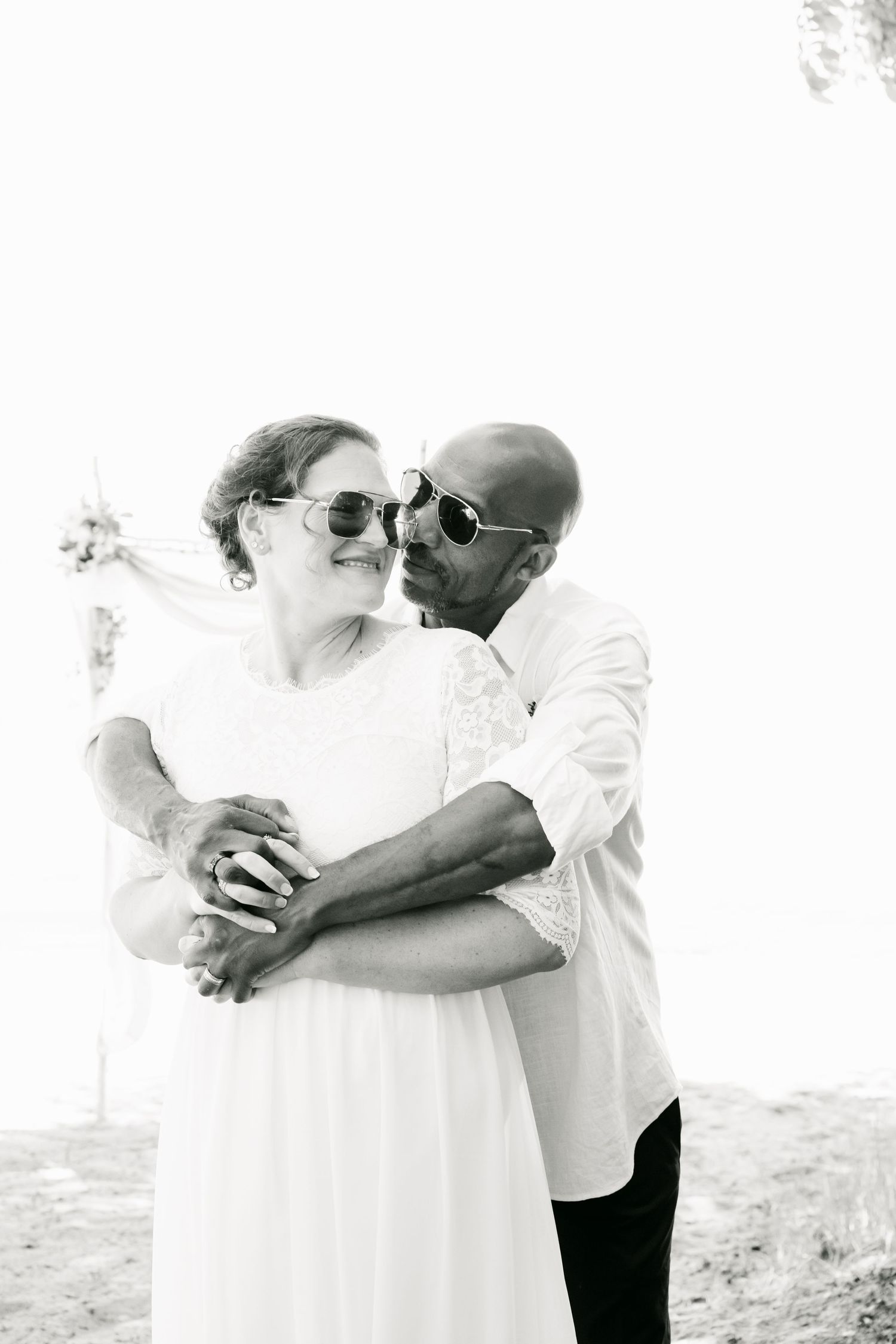 Black and white photo of interracial couple embracing in sunglasses at outdoor wedding.