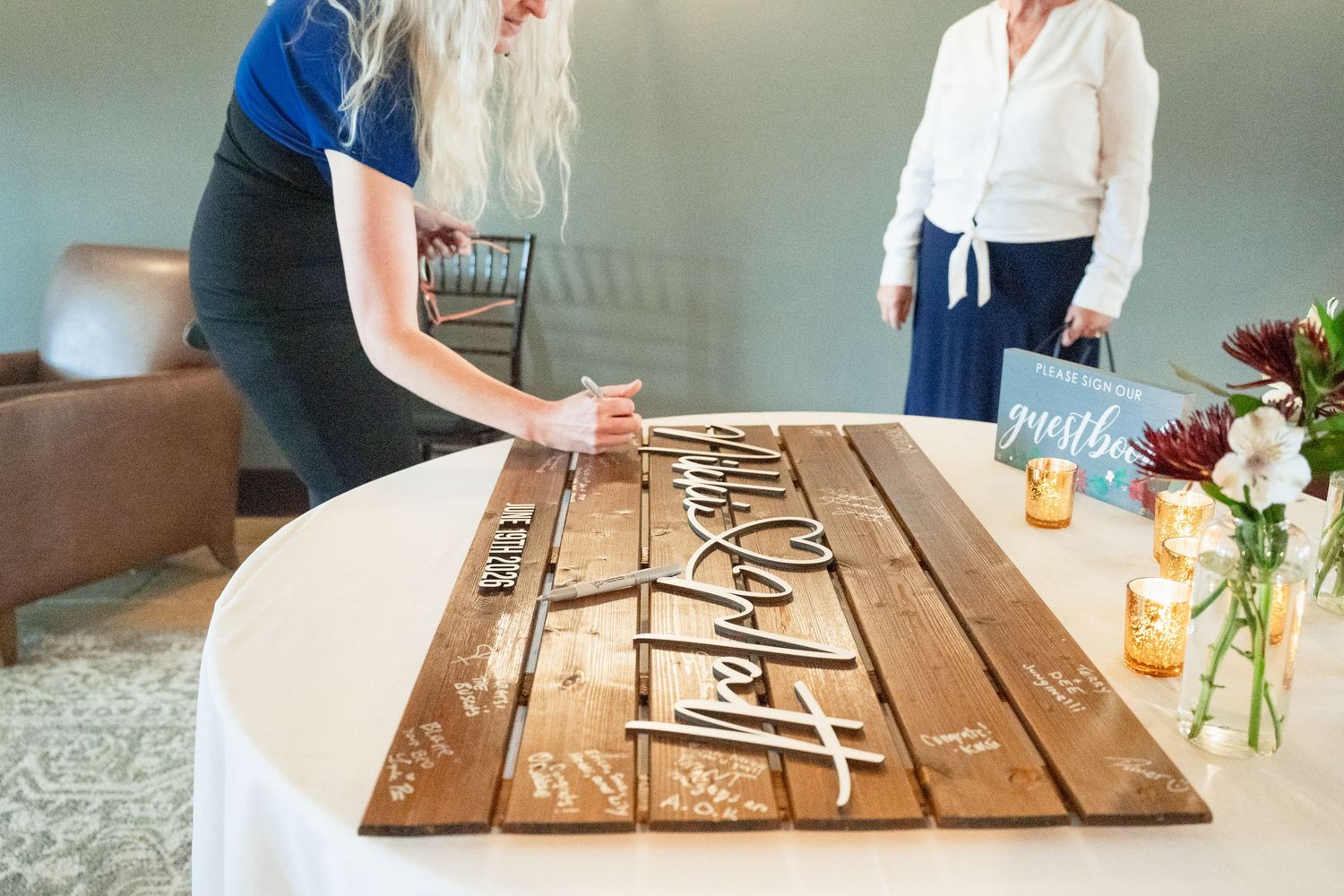 Guests sign a rustic wooden wedding guest book with elegant calligraphy at a decorated reception table.