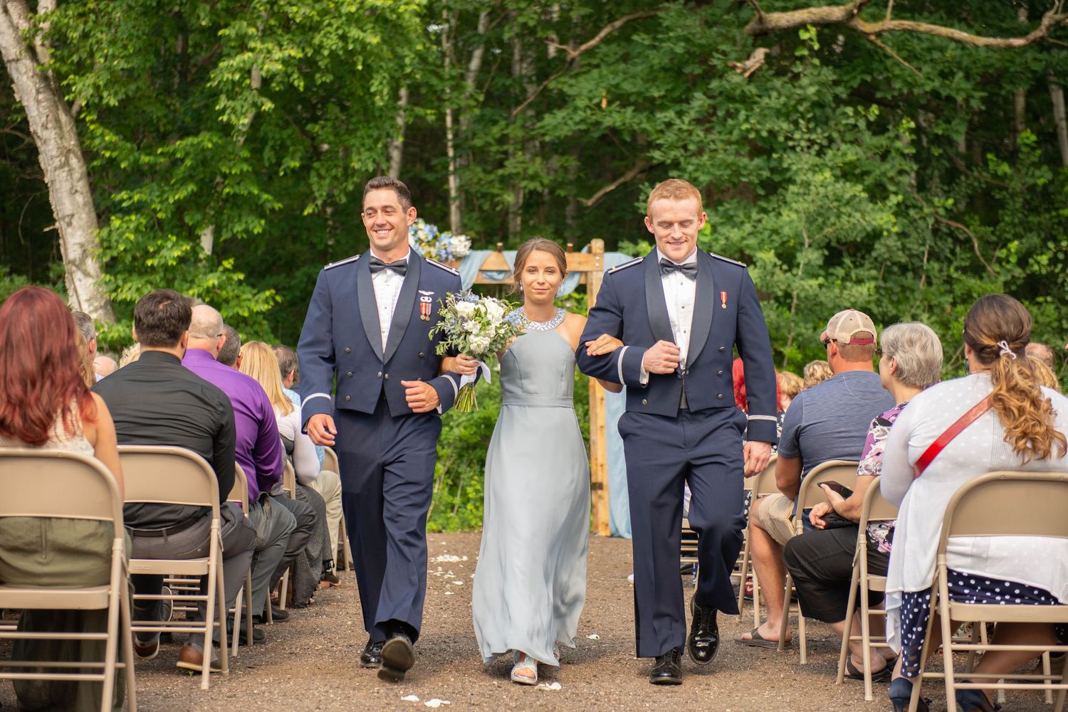 Three wedding attendees walk down an outdoor ceremony aisle between rows of folding chairs in a wooded setting. Zimmerman MN Weddings