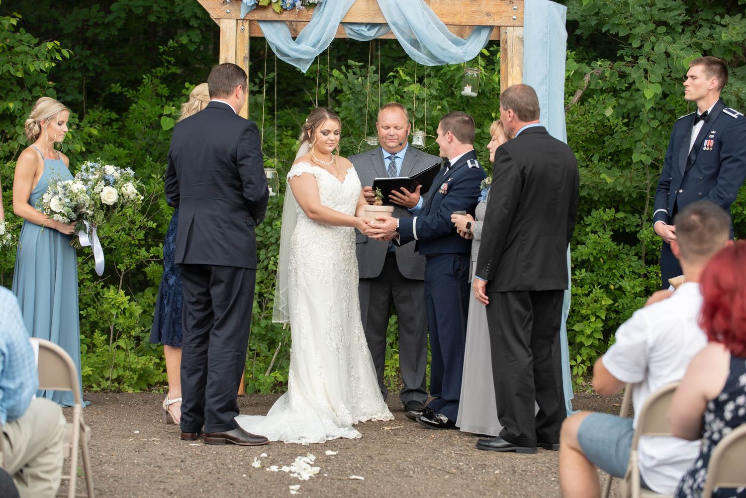 Outdoor wedding ceremony under wooden arch with light blue drapery and greenery backdrop. Zimmerman MN Weddings