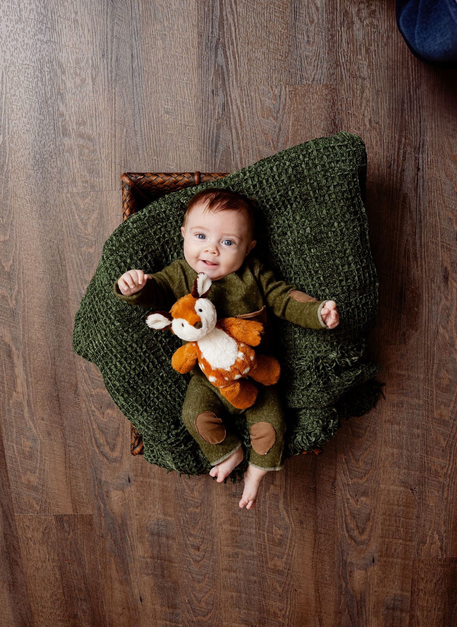 Baby wearing brown outfit lies on green knit blanket holding orange fox plush toy on wooden floor. Princeton MN Milestone, Maternity and New