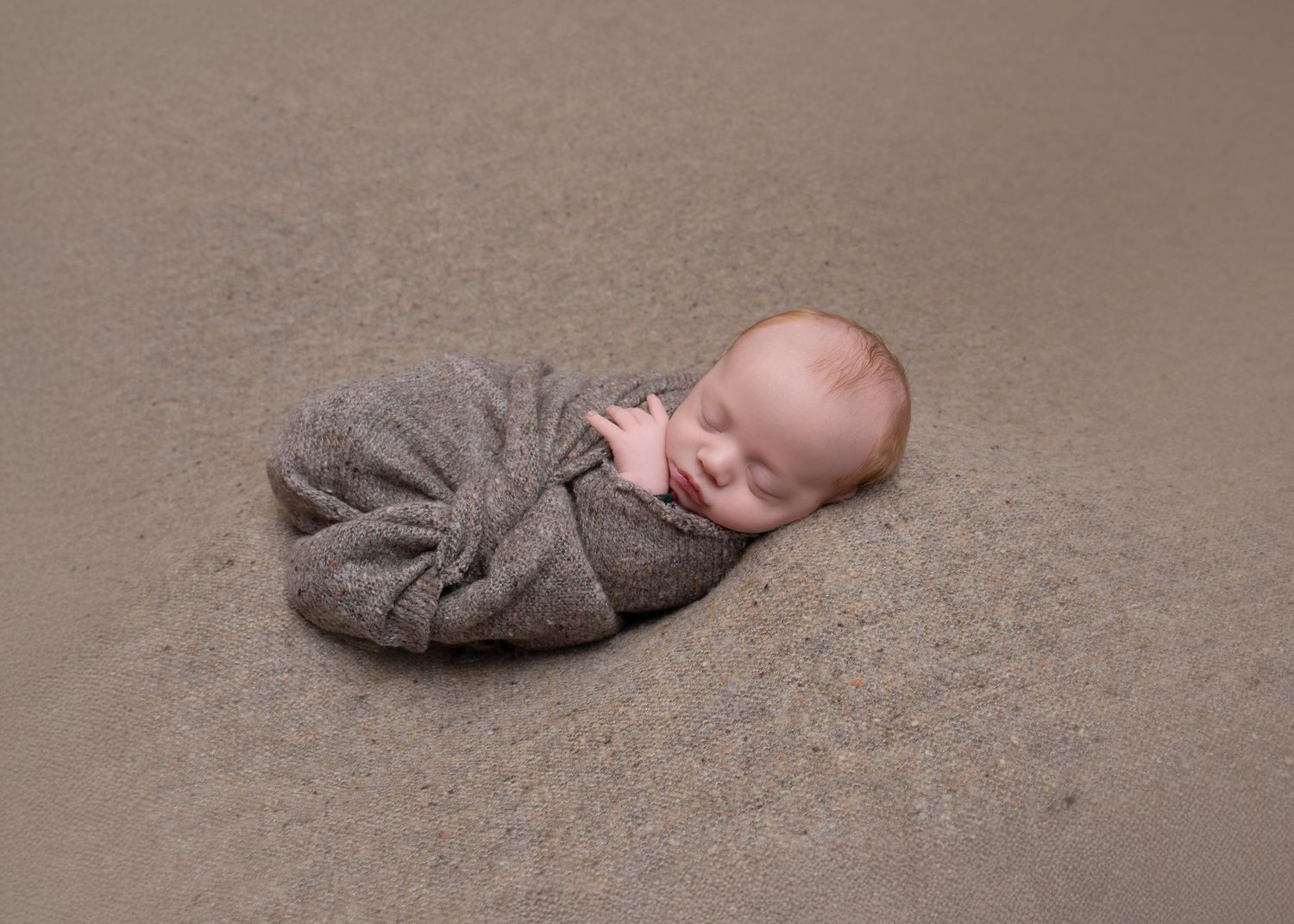 Newborn baby peacefully sleeping while wrapped in soft gray blanket on tan backdrop.