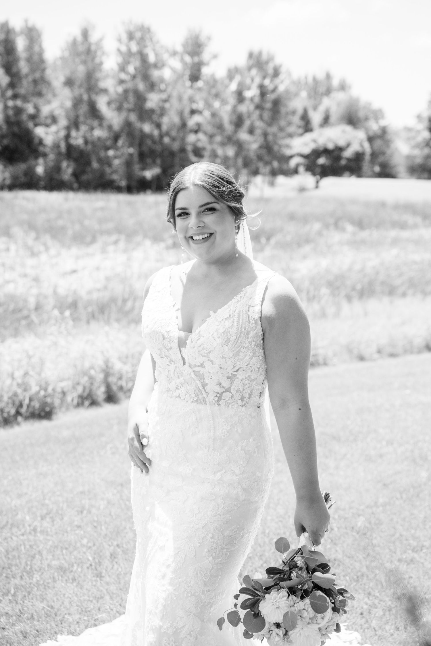 Elegant bride in lace wedding gown holding bouquet poses outdoors in black and white portrait.