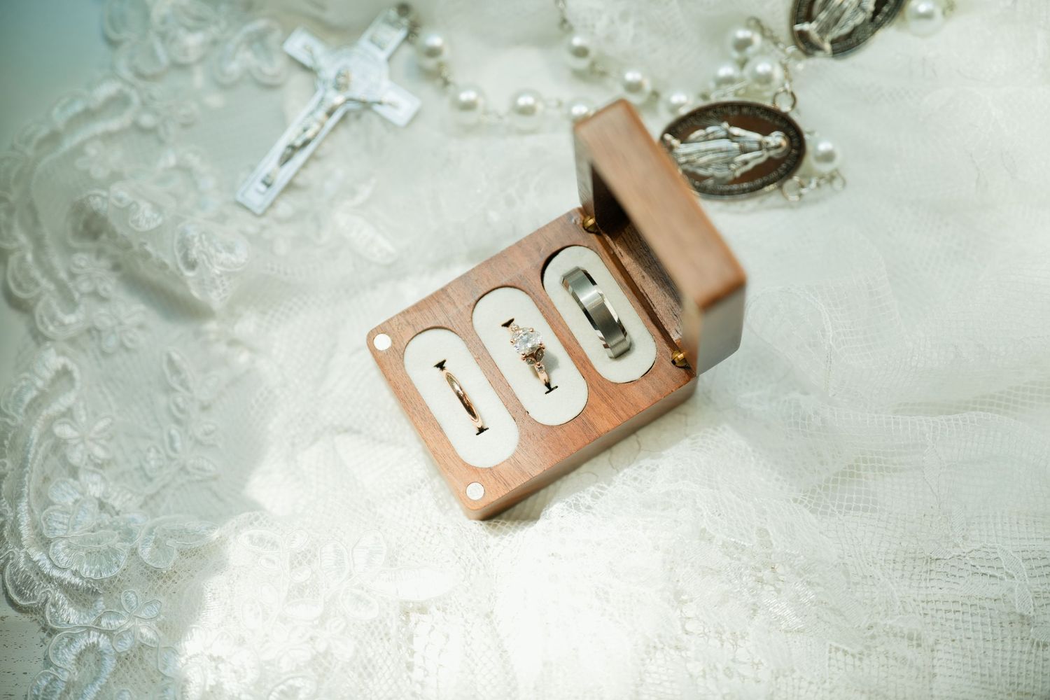 Wooden ring box with three slots holding wedding bands, placed on white lace fabric with rosary beads nearby.