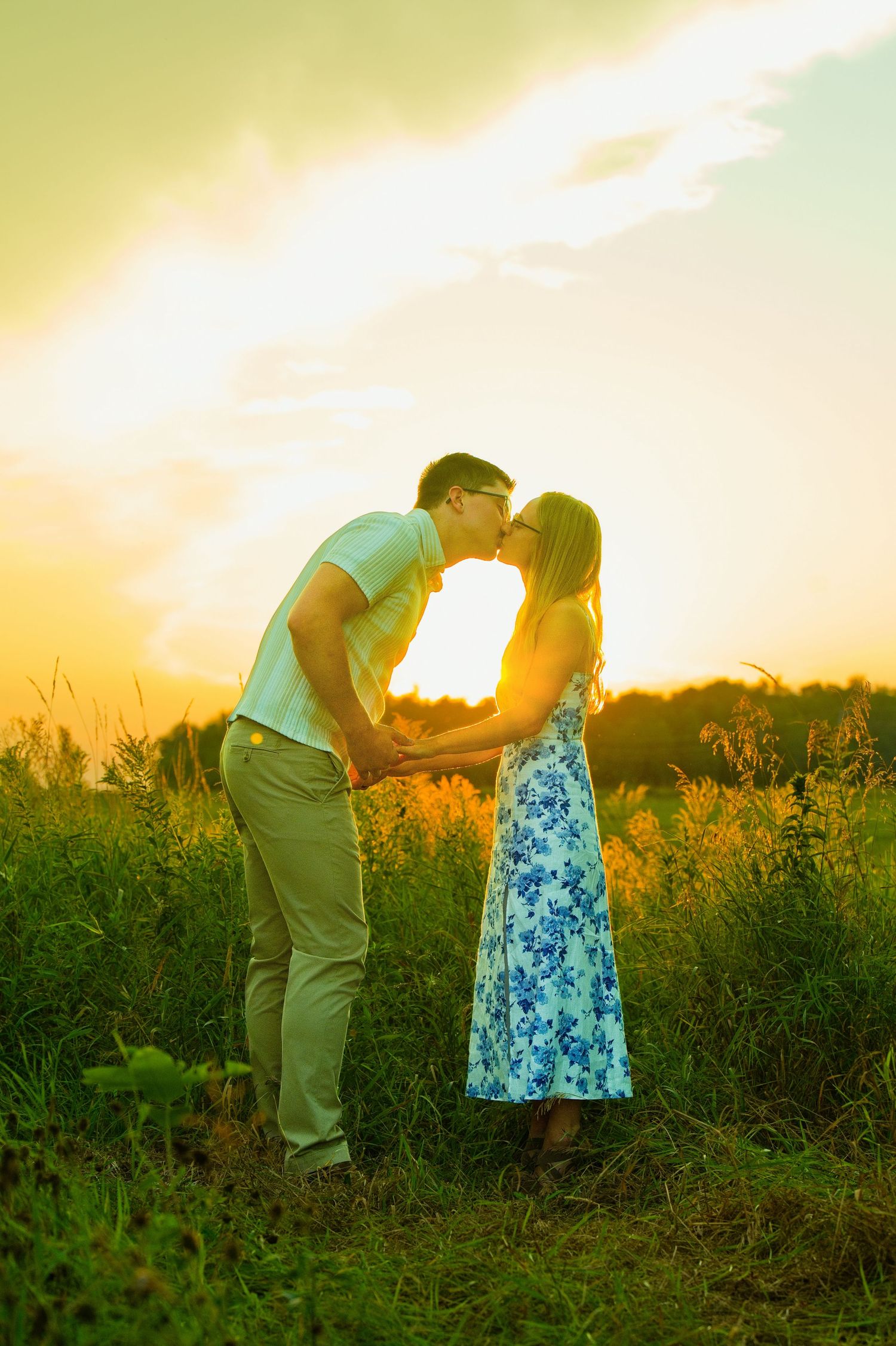 Couple sharing a romantic kiss at golden sunset in a lush green field, woman in floral dress holding hands with man.