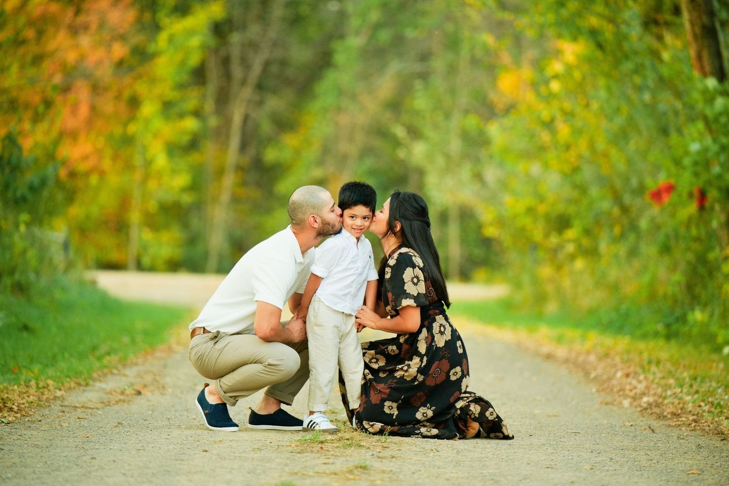 Parents kissing their young son on the cheeks on a scenic tree-lined path during autumn family photo session.