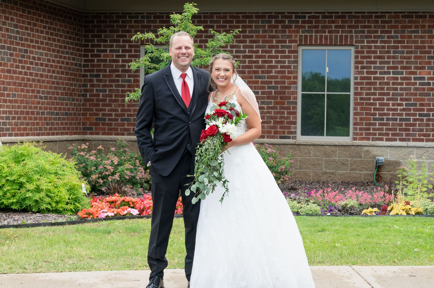 Bride and groom pose together outside a brick church, bride holding a cascading red and white bouquet.