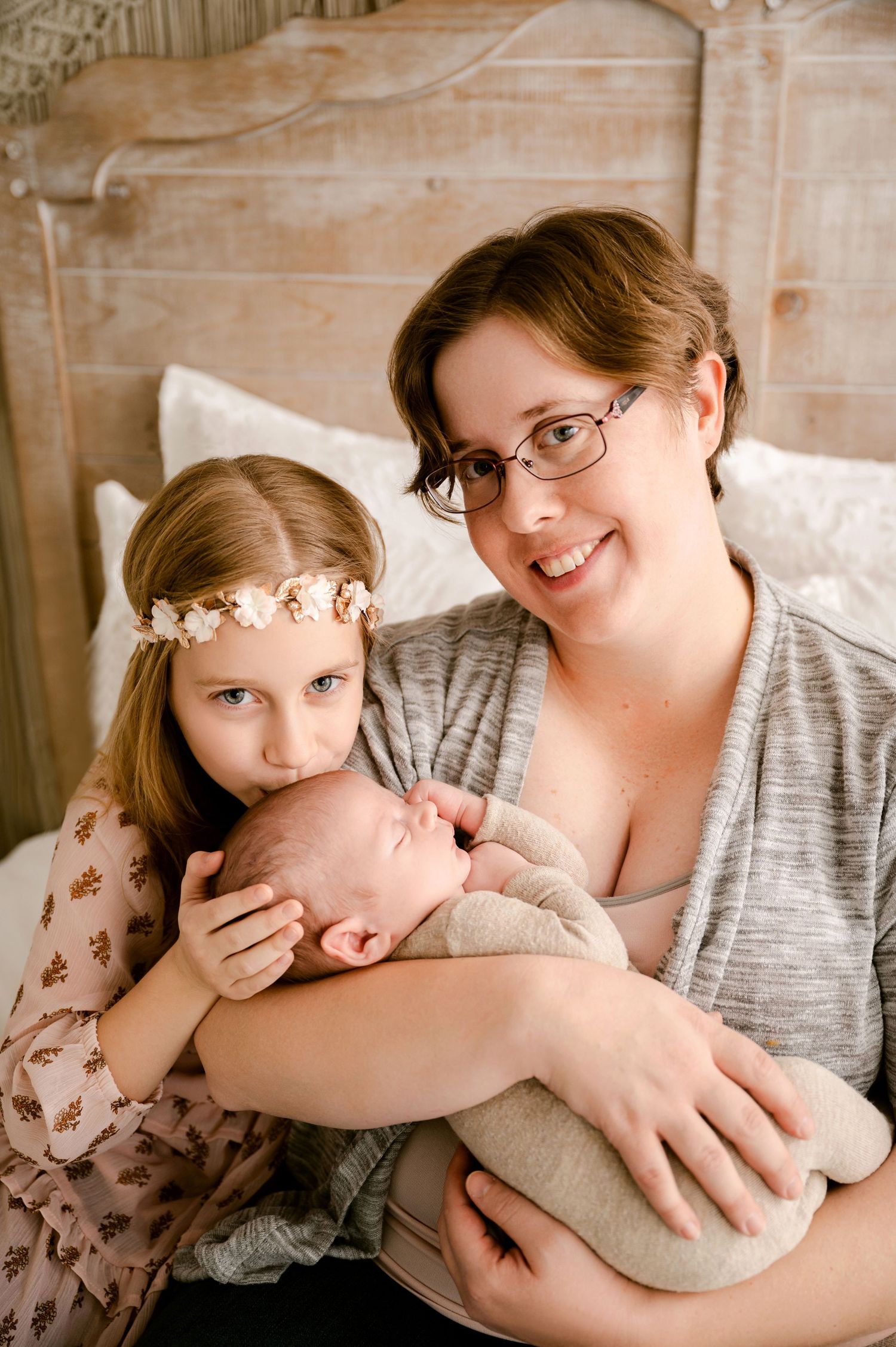 Indoor family portrait shows mother and children embracing together on bed with rustic wooden headboard.