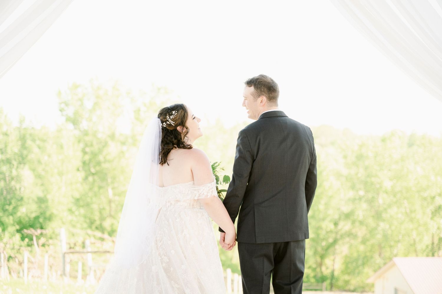 Central MN Wedding Photos - Bride and groom hold hands under white draped arch, surrounded by lush greenery on their wedding day.