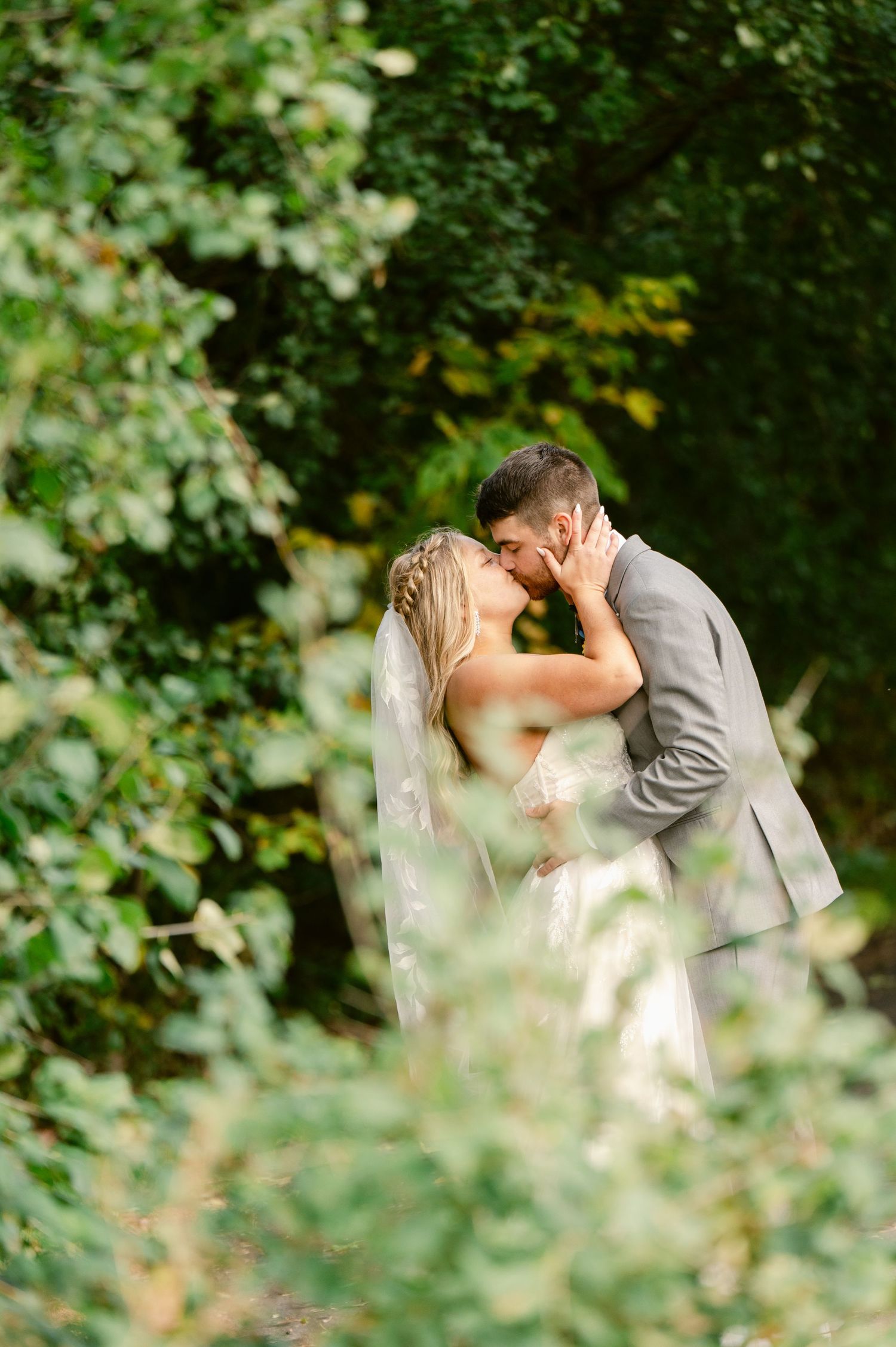 Central MN Wedding Photos | Bride and groom kissing softly surrounded by lush green trees in an ethereal outdoor wedding portrait.