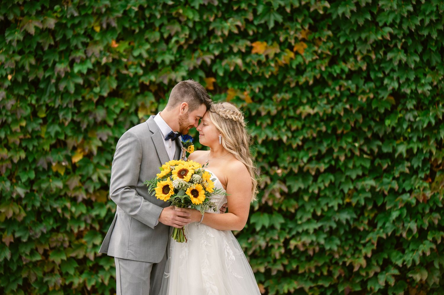 Central MN Wedding Photos | Bride and groom share a romantic moment holding sunflower bouquet against lush green ivy wall.