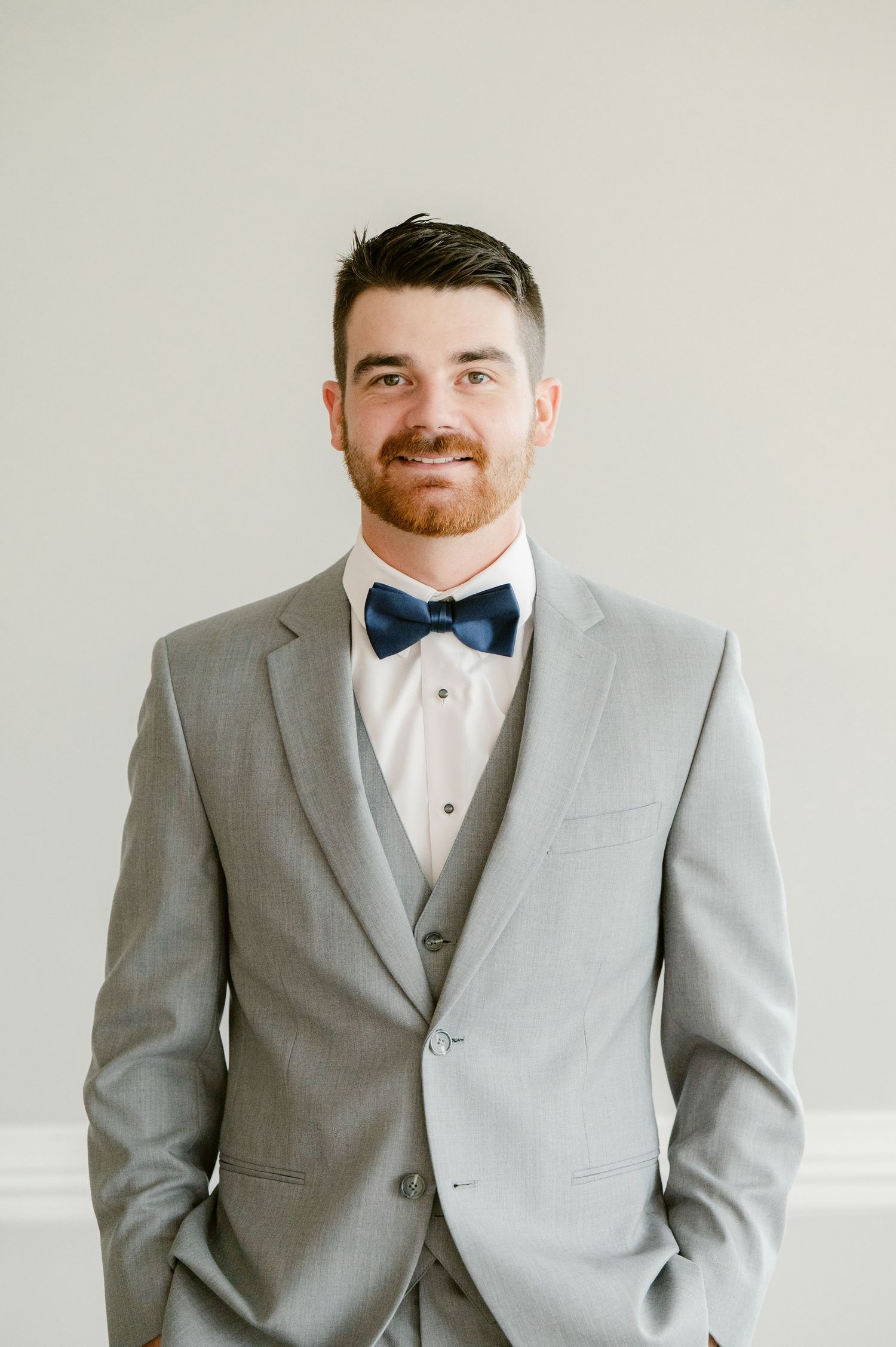 Central MN Wedding Photos | Handsome groom in gray three-piece suit and navy bow tie smiles confidently against white wall.