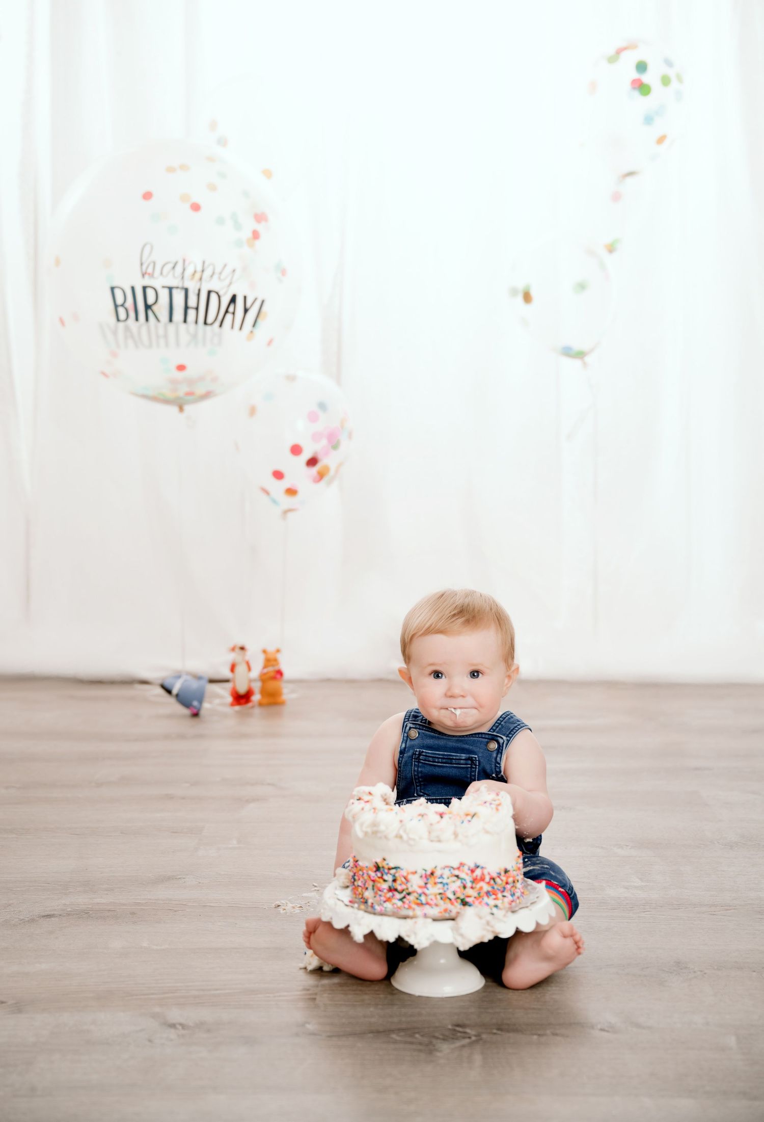Central MN Photos - Baby boy in denim overalls sitting on floor with birthday cake and colorful balloons during first birthday cake smash.