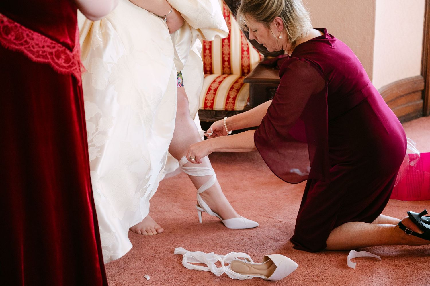 Central MN Wedding Photos | Bridesmaid helping bride put on white heels before wedding ceremony.
