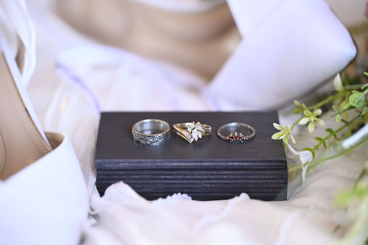 Close-up of wedding rings displayed on a dark wooden box with delicate white flowers and greenery.