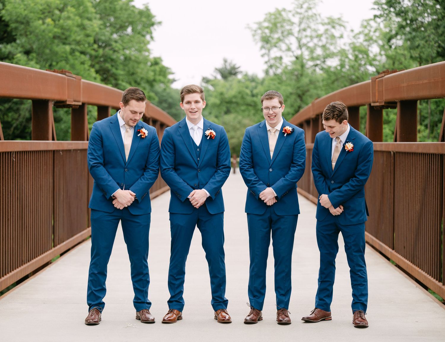 Four groomsmen in matching navy blue suits and boutonnieres stand together on a wooden bridge outdoors.