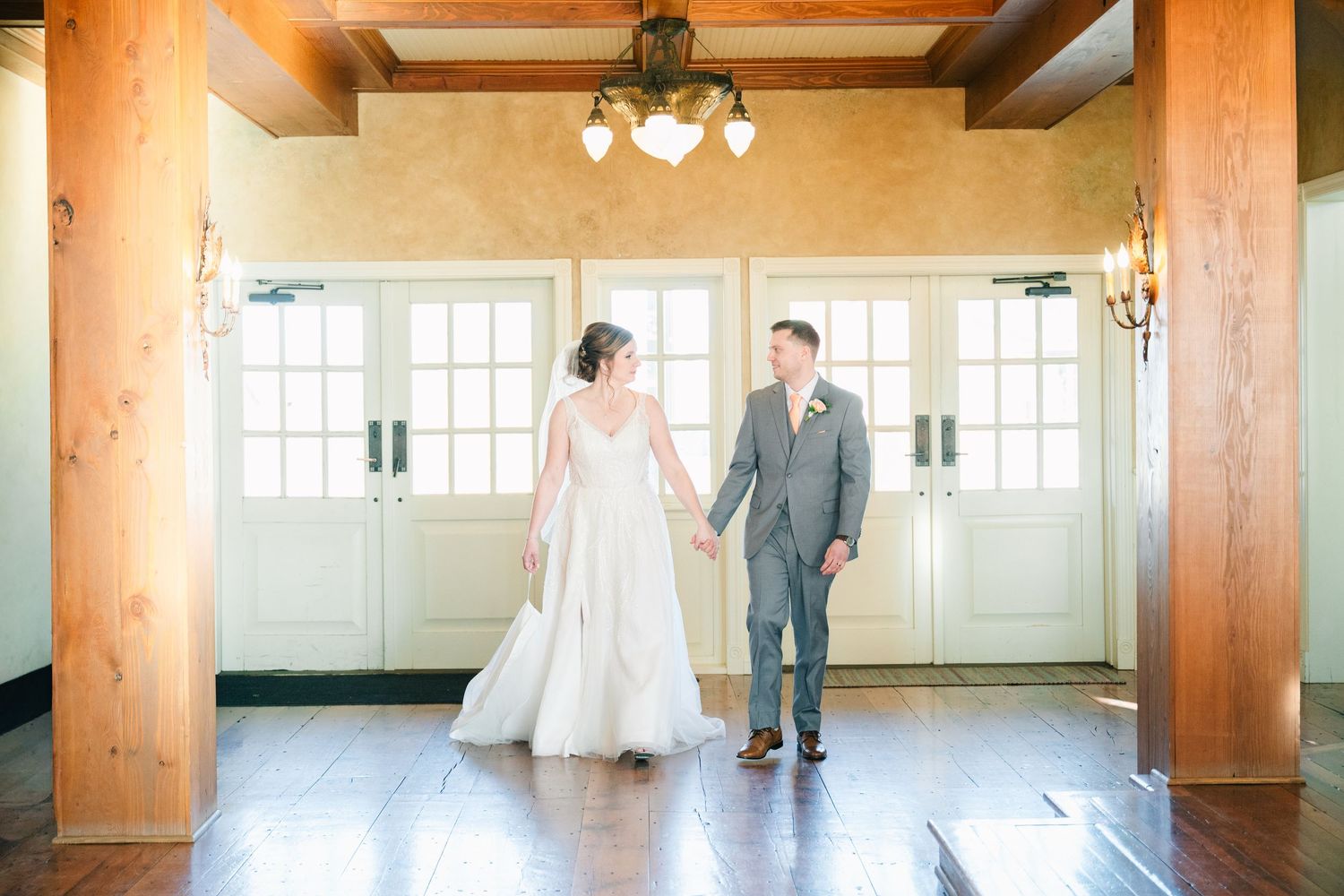 Bride and groom holding hands smiling in elegant venue with French doors and chandelier.