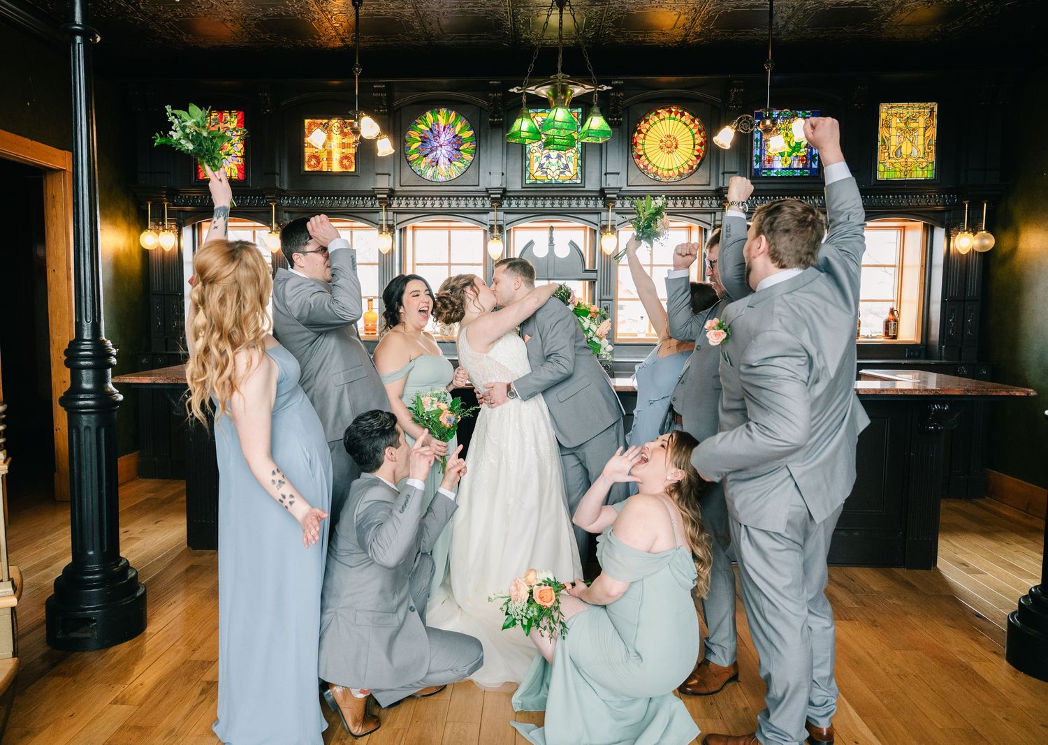 Wedding party celebrating newlyweds with a joyful group hug inside a rustic bar venue with stained glass windows.
