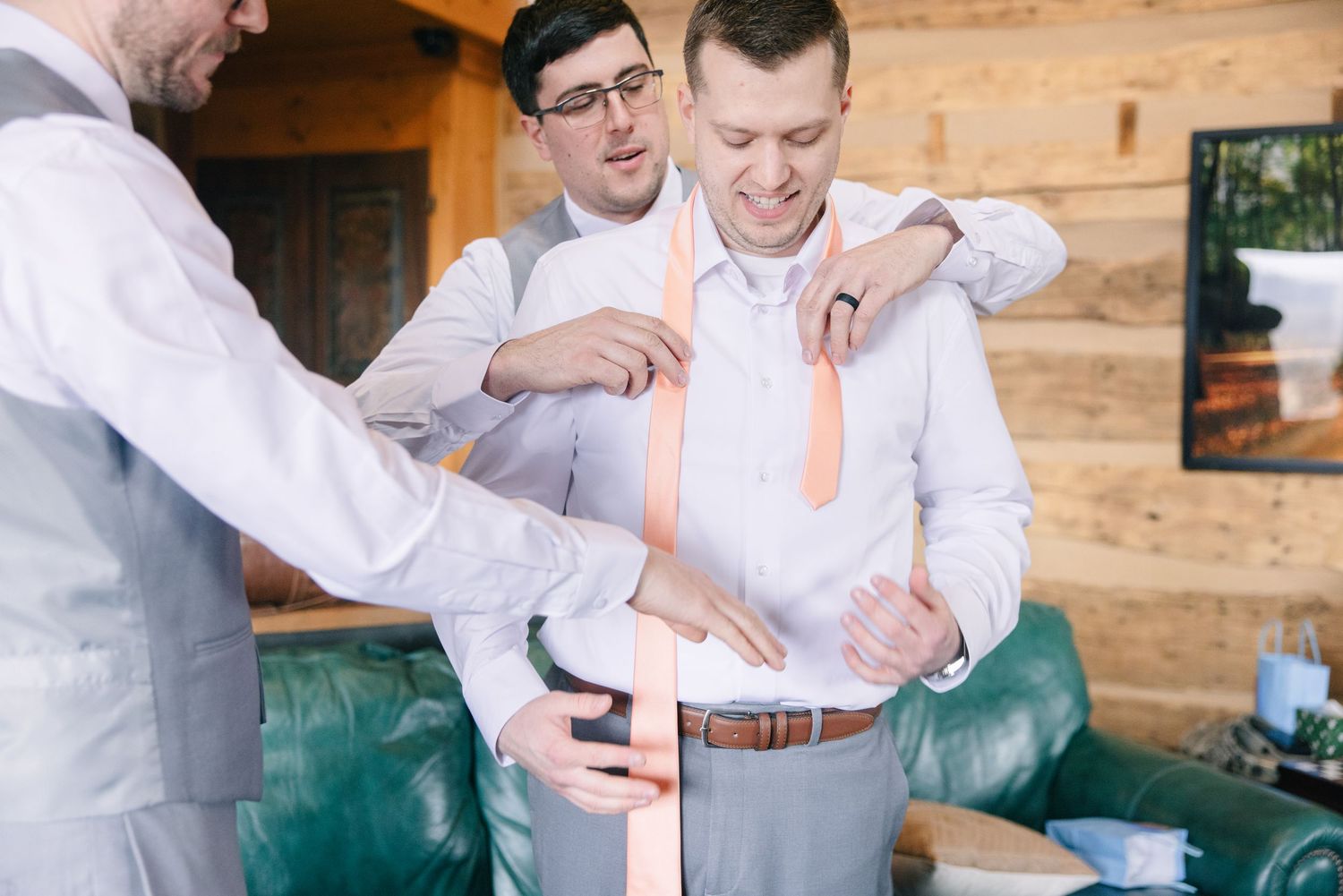 Groomsmen helping groom adjust his peach tie and suspenders while getting ready in a rustic cabin.