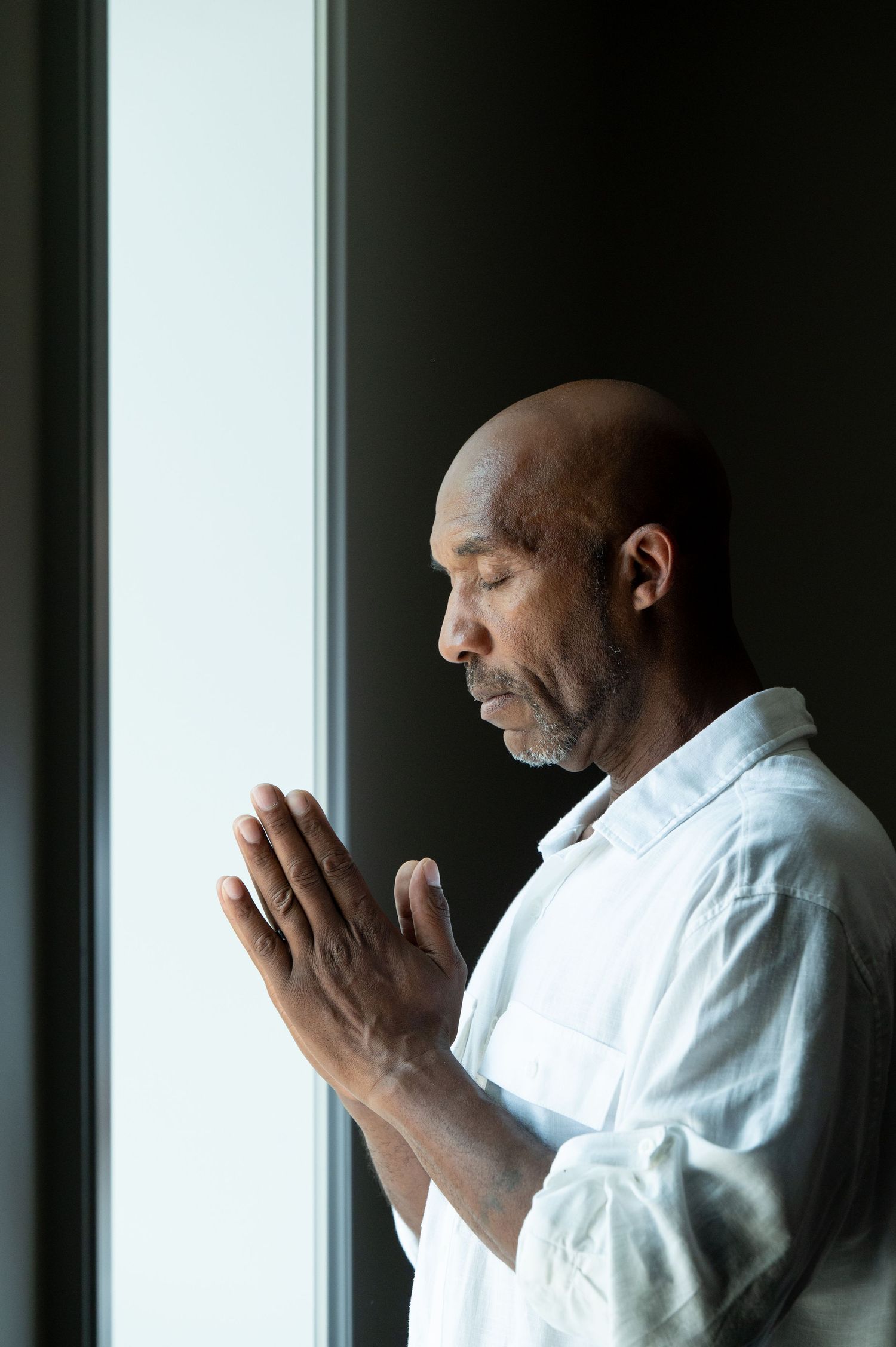 Bald middle-aged Black man in white shirt praying with hands pressed together near a window.