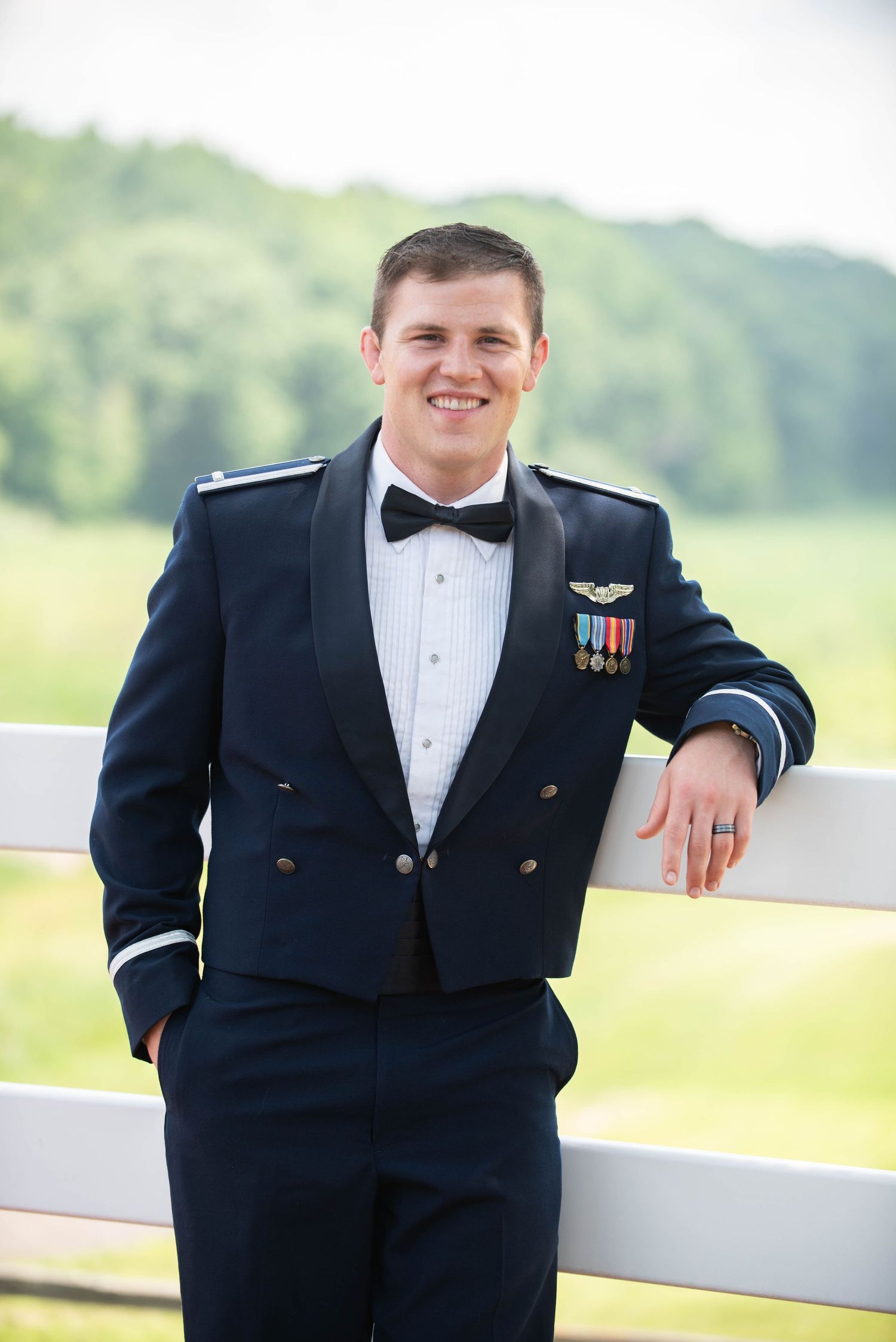 Military groom in dress uniform with medals leaning against white fence rail. Zimmerman MN Weddings