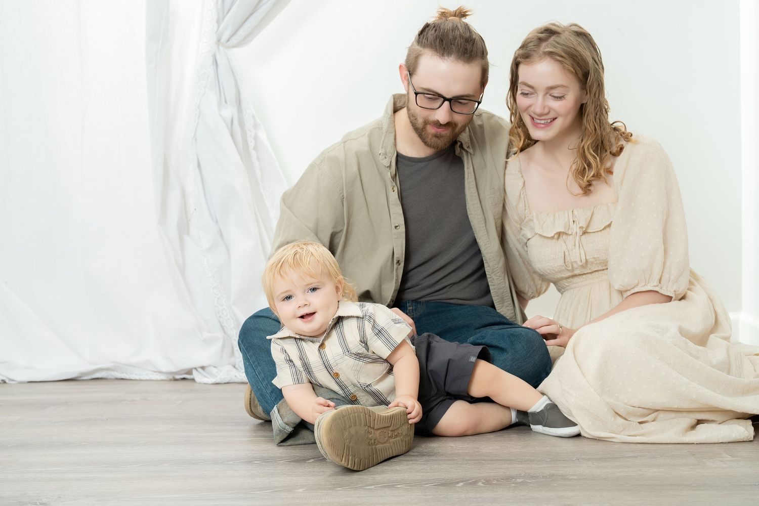 A young family sits together on a light wooden floor against a white backdrop during a casual portrait session. Princeton MN Milestone, Mate