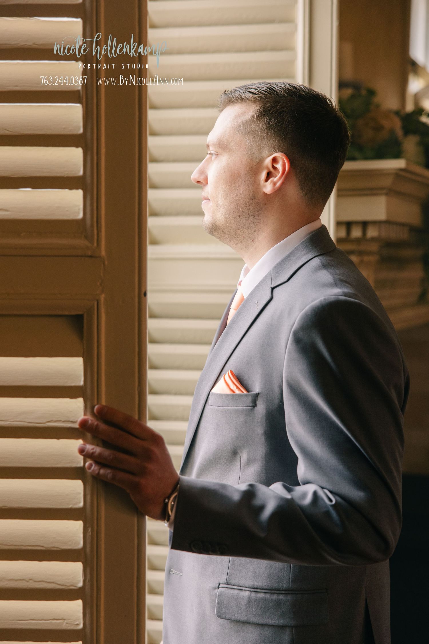 Groom in gray suit with pink pocket square gazes thoughtfully out window through wooden shutters on wedding day. Crooked Willow Osakis