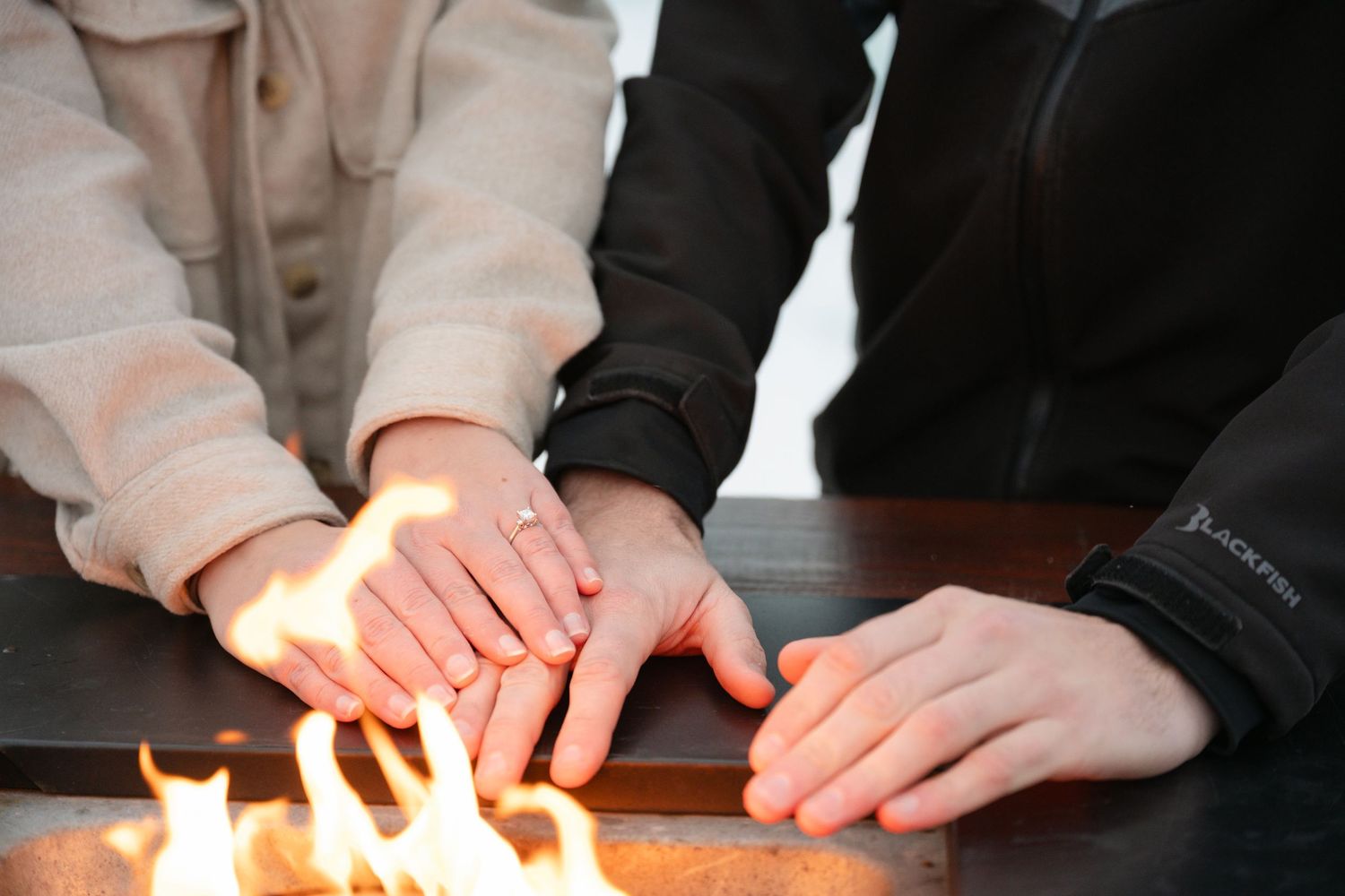 Two people warming their hands over an open flame fire pit on a cold winter day outdoors. Engagement Minneapolis MN