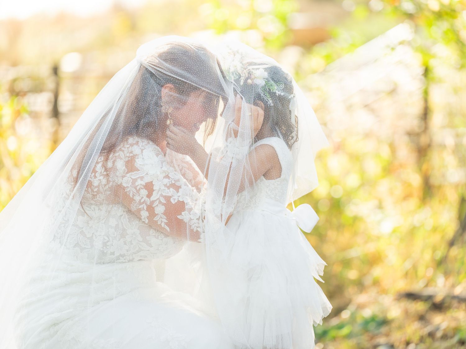 Bride and flower girl share a tender moment under a flowing veil in a sunlit outdoor wedding setting.