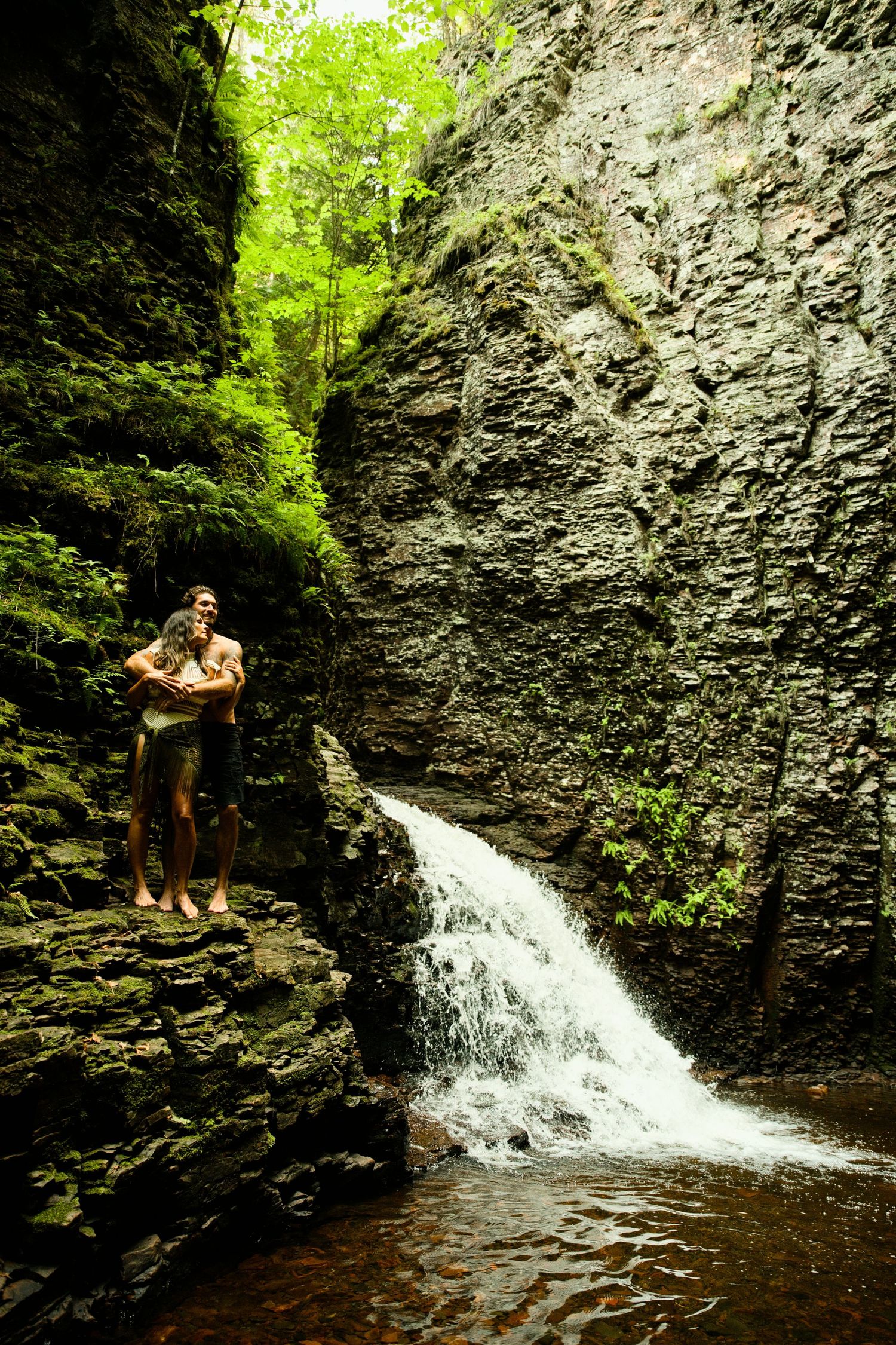 Couple embracing on rocks beside a cascading waterfall in a lush green canyon.