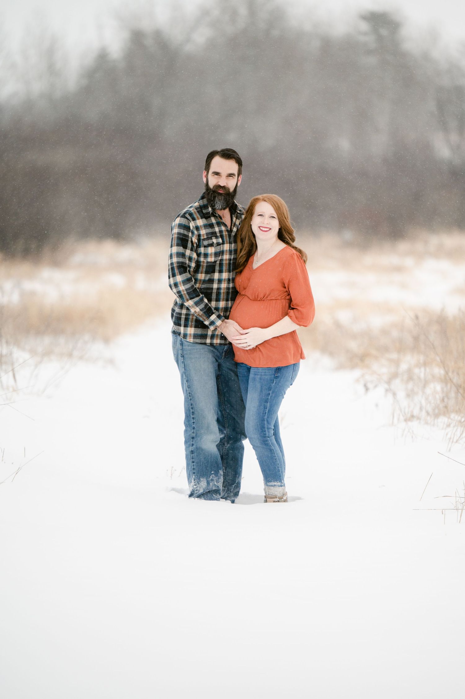 The Clark’s romantic winter maternity photoshoot together in a snowy winter field, wearing casual clothing of plaid and orange sweater.