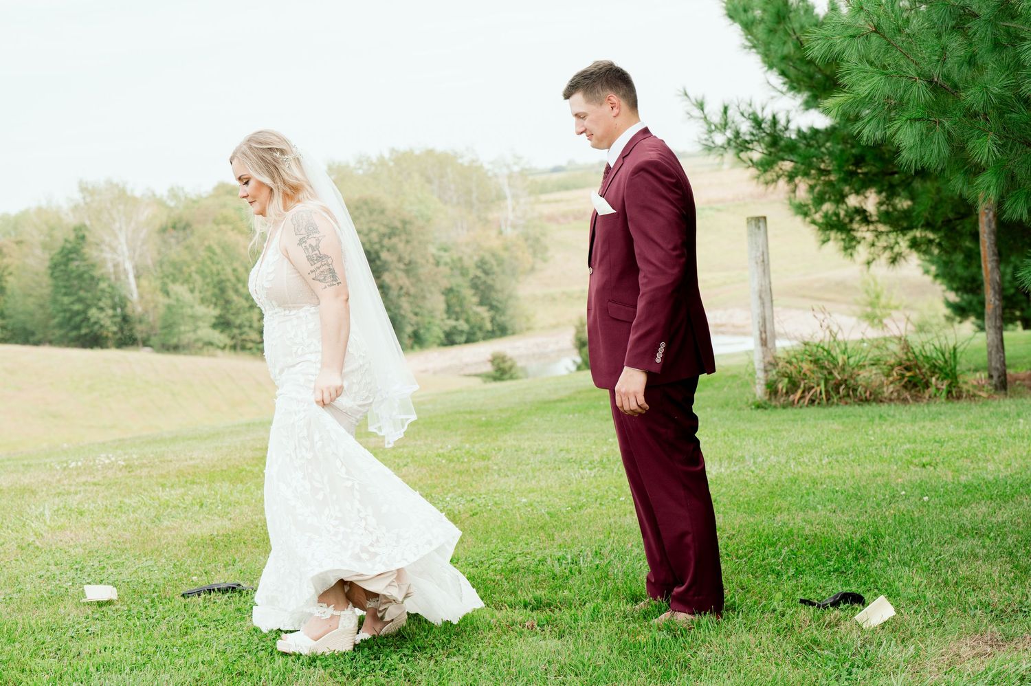 Newlyweds share a playful moment standing back-to-back on a grassy hill during their wedding photoshoot.