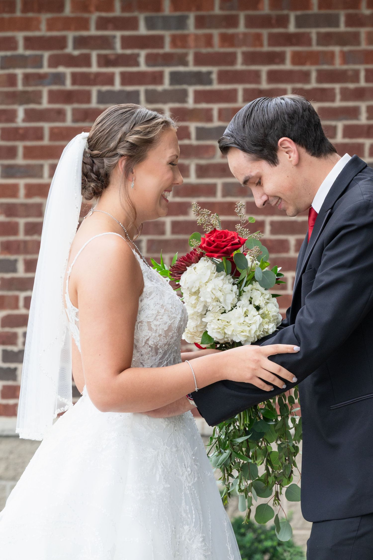 Bride and groom share a joyful first look moment, holding a red and white bouquet against a brick wall.