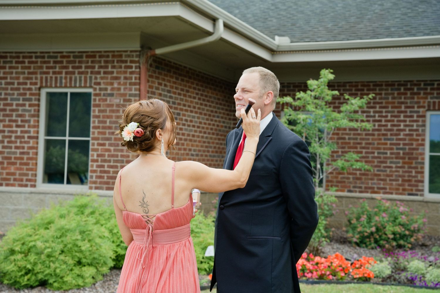 Couple in formal attire outside brick building, woman in coral gown touches man's face as he talks on phone.