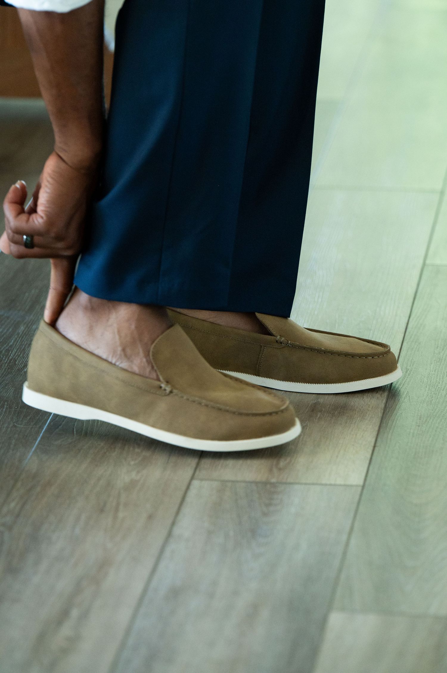 Man wearing tan suede loafers with white soles and navy dress pants standing on hardwood floor.