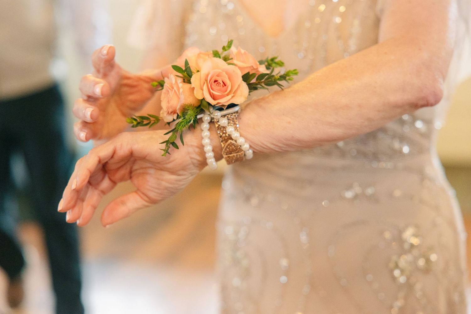 Close-up of wrist corsage with peach roses and pearl bracelet on wedding guest in beige dress.