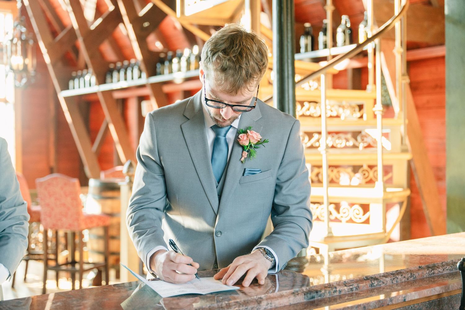 Groom in gray suit signing wedding documents at a wooden bar in a rustic venue.