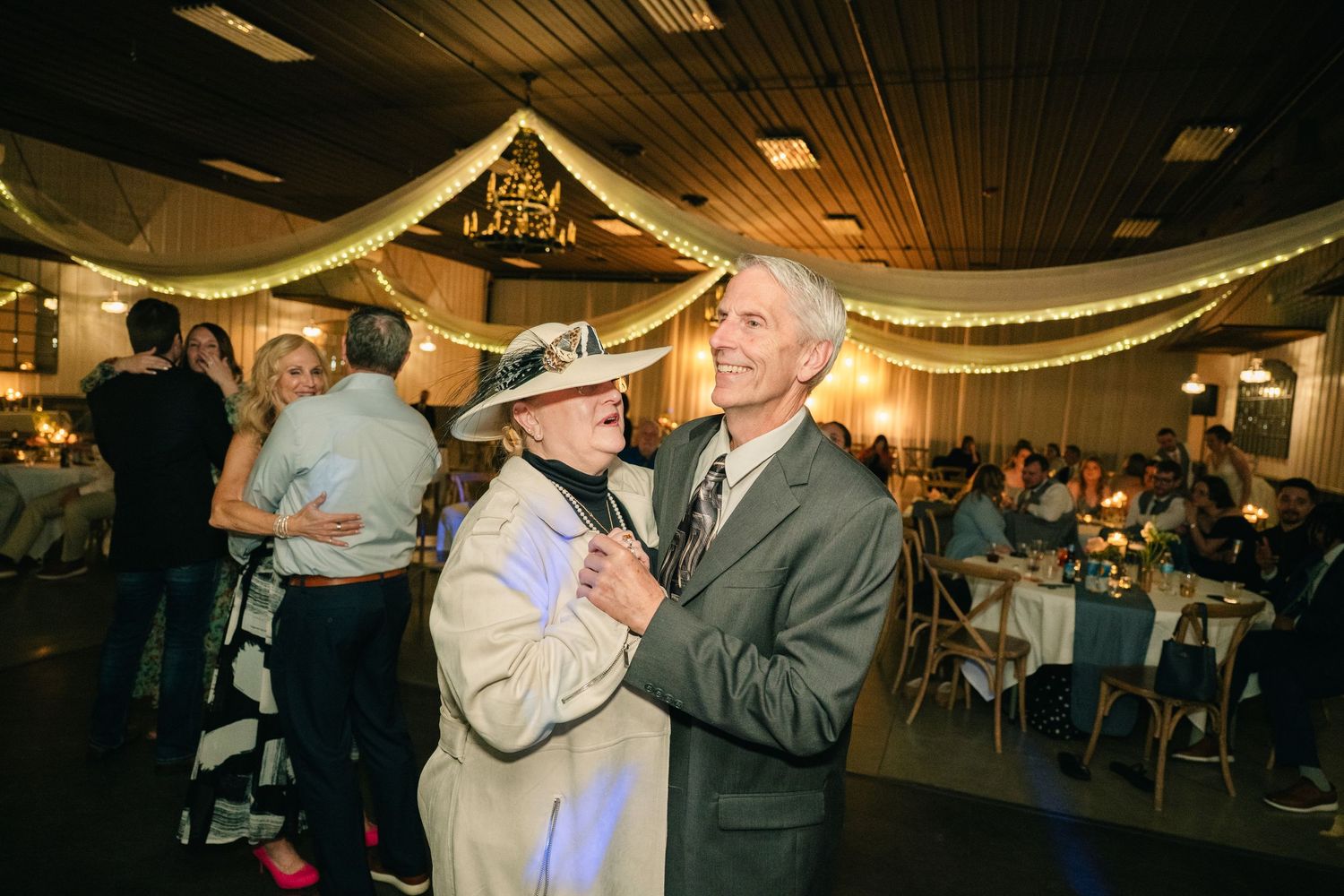 Elderly couple dancing joyfully at a wedding reception under warm string lights.