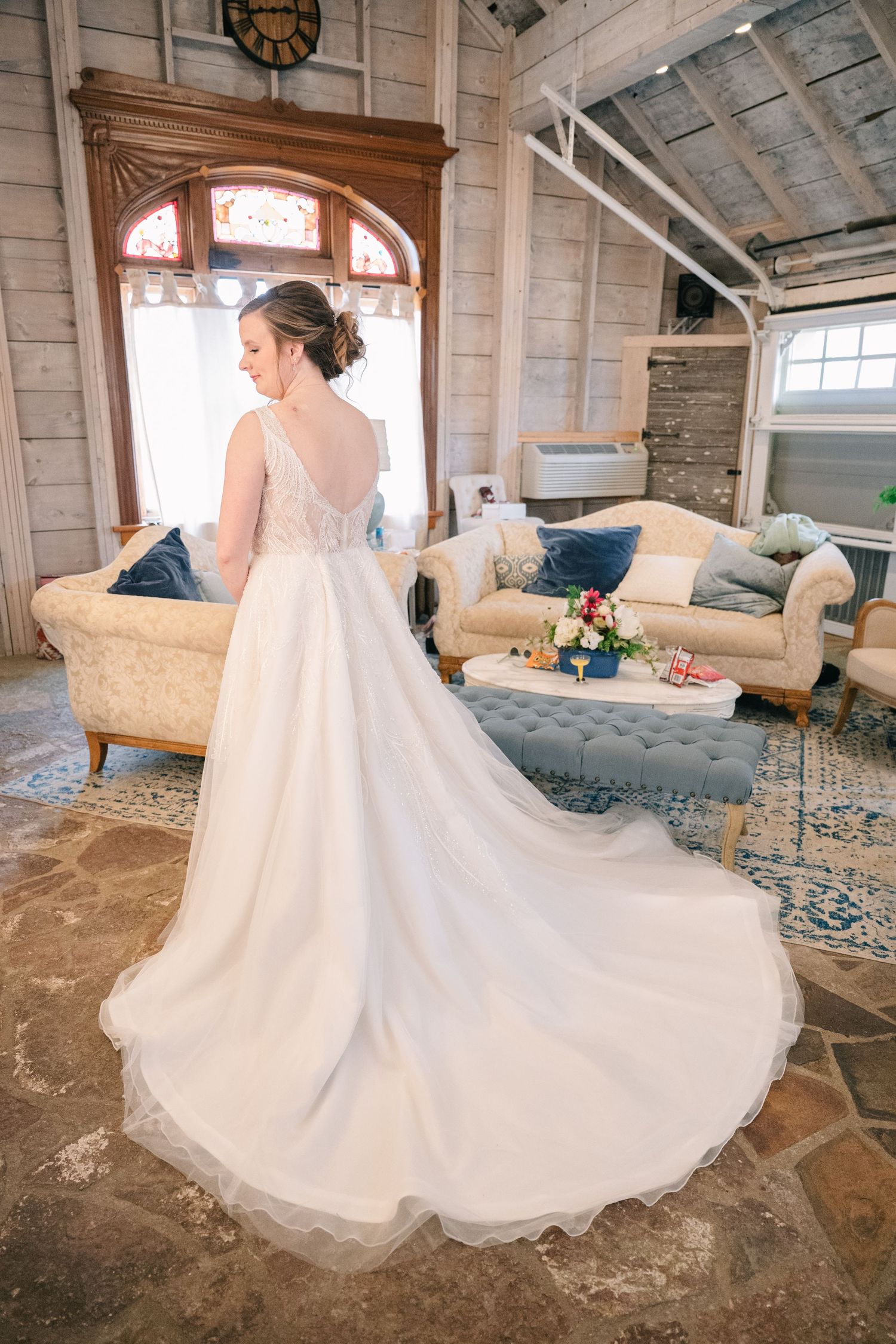 Bride in a stunning backless white ball gown with long train standing in a rustic venue with vintage furniture.
