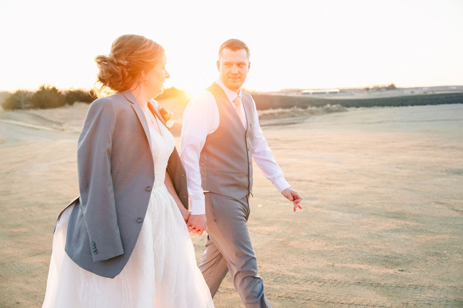 Bride and groom holding hands walking on sandy dunes during golden sunset after wedding ceremony.