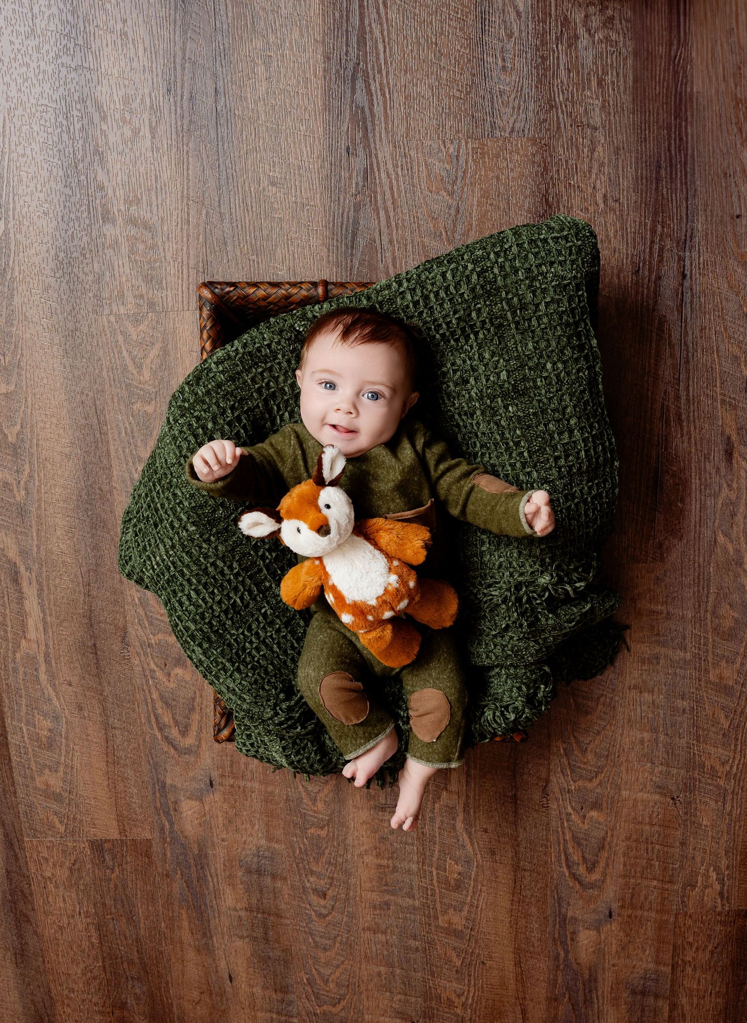 Central MN Milestone Photography - Baby in green outfit lying in a basket with a stuffed fox toy on a wooden floor.