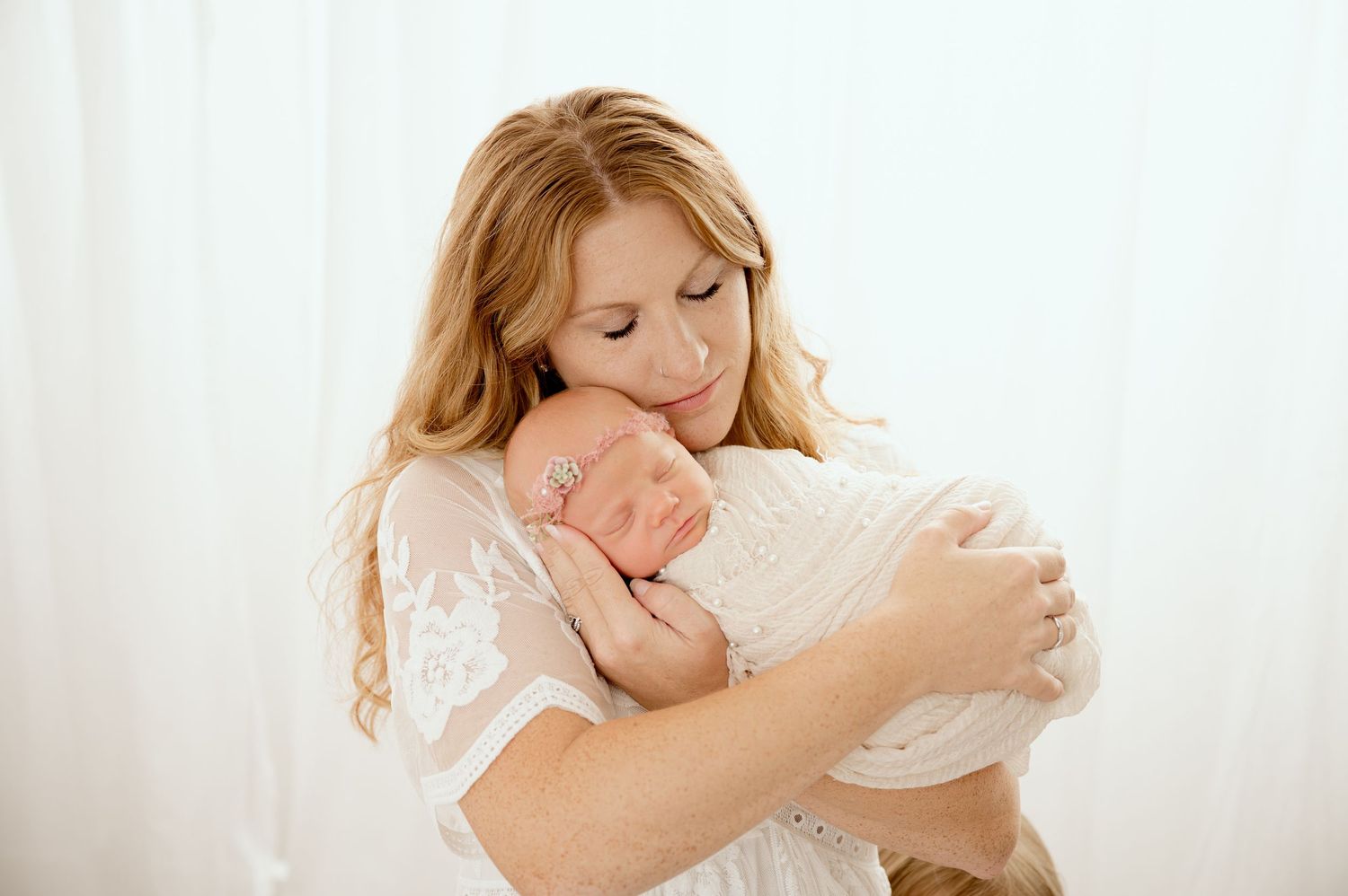 Smiling blonde mother in white lace dress gently cradles her sleeping newborn baby close to her face.
